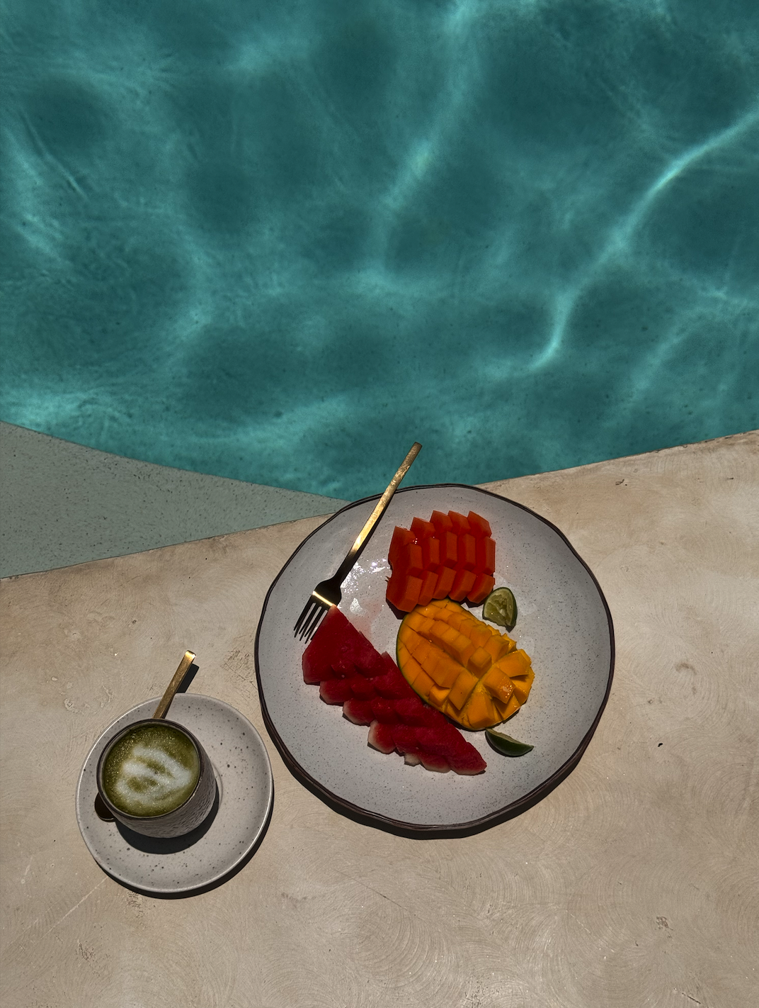 Assorted sliced fruits including watermelon, cantaloupe, honeydew, and lime on a plate, with a small cup of green smoothie nearby, set near a swimming pool.