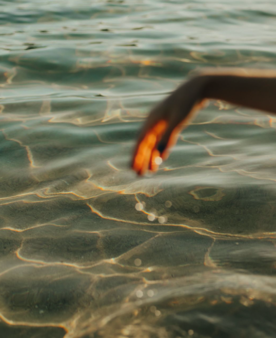 A person dipping their hand into clear, rippling water at sunset.
