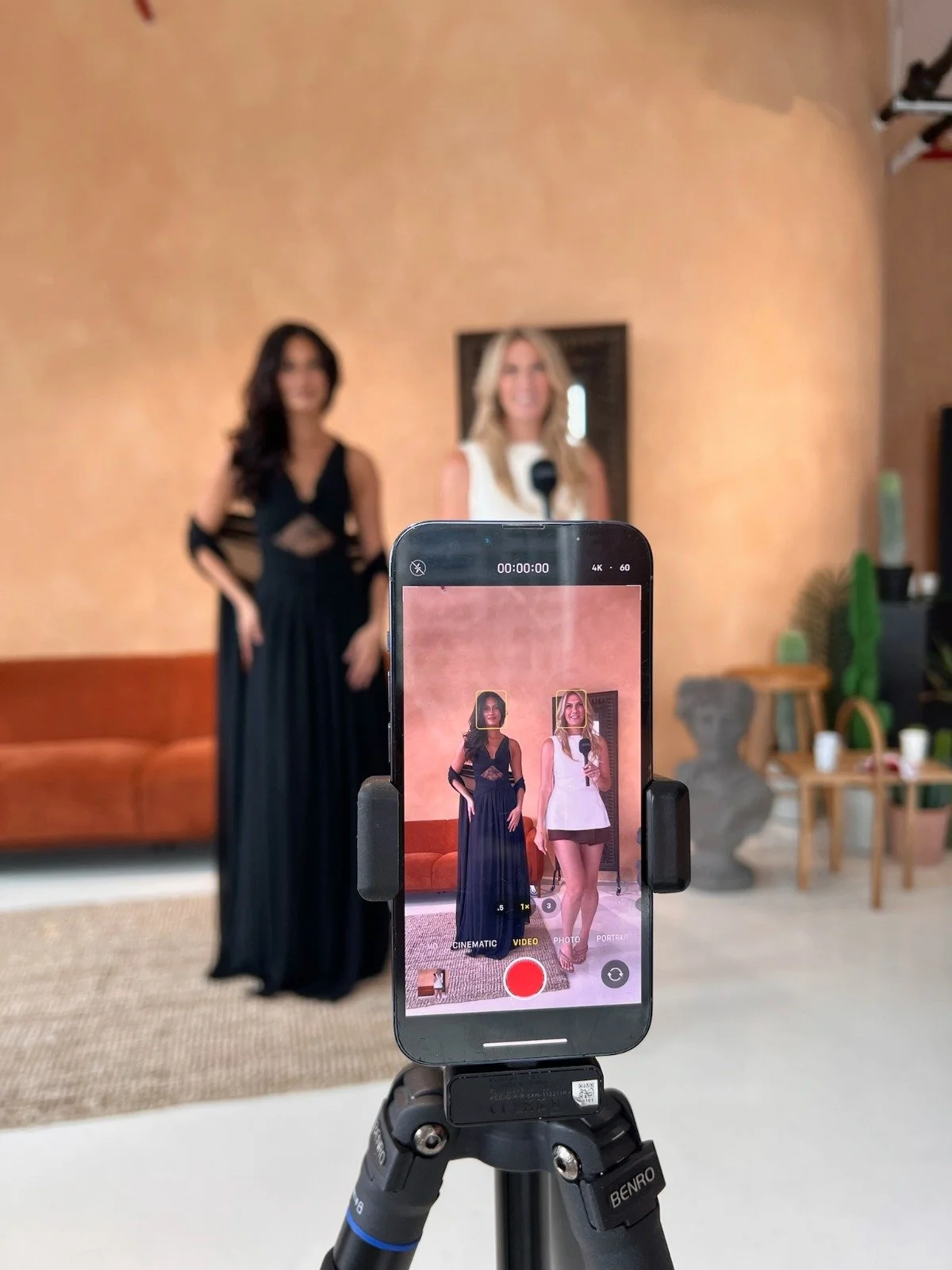 A smartphone on a tripod recording two women standing in front of a beige wall, with one woman in a black dress and the other in a white dress holding a microphone.