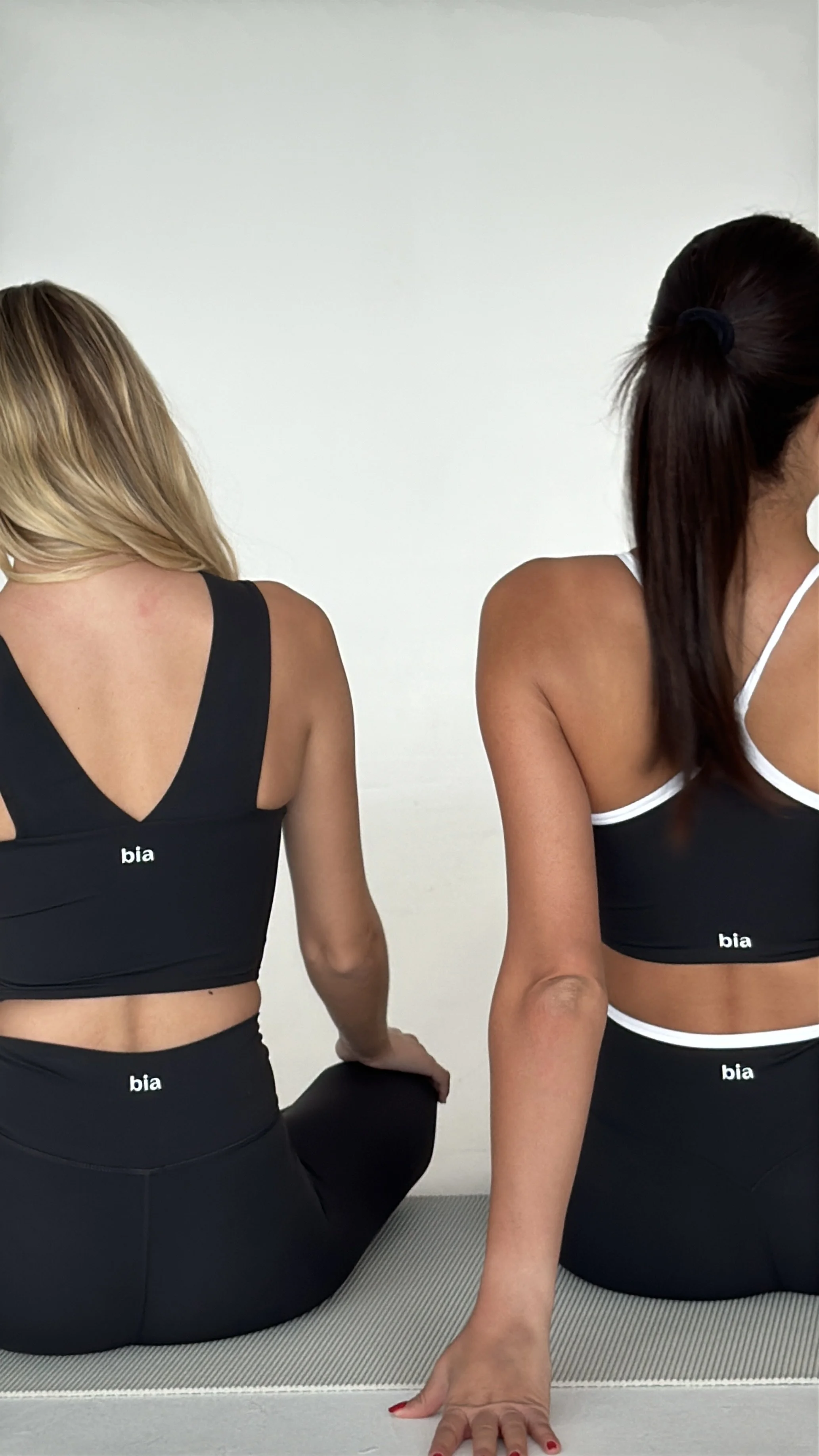 Two women sitting on yoga mats, practicing yoga or meditation. They are wearing matching black sports bras with white straps and the word 'bia' printed on the back. One woman with blond hair is sitting with her legs crossed, and the other woman with dark hair tied back is sitting upright with her left hand on the floor.