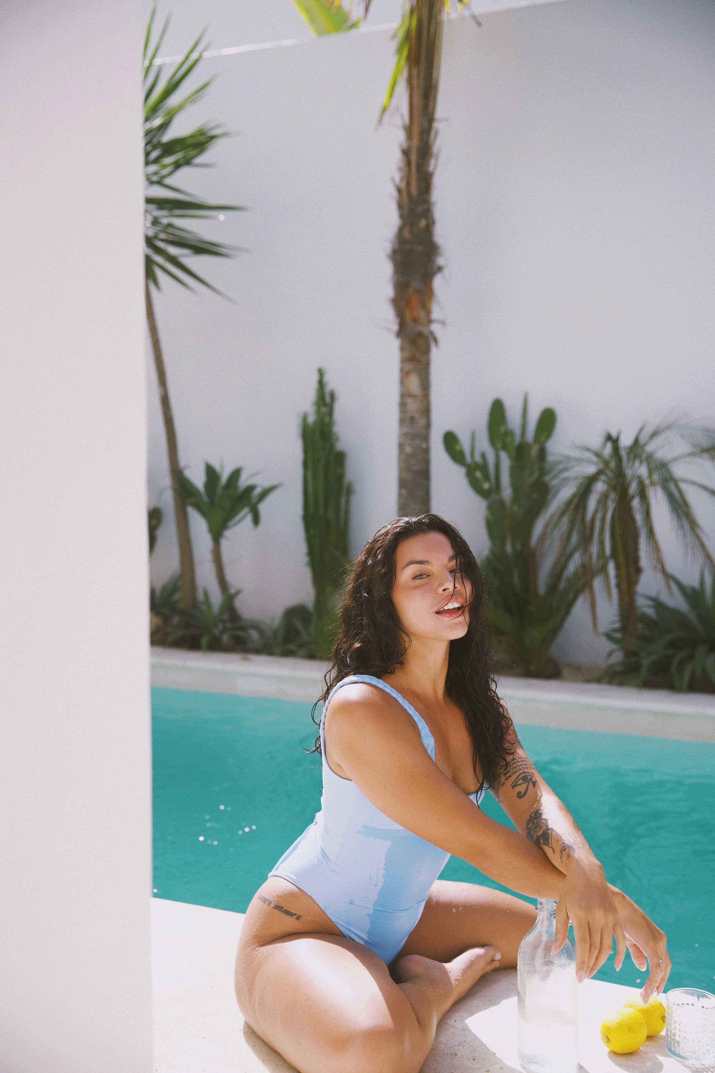 A woman with long curly hair, wearing a light blue swimsuit, sitting by a pool, holding a glass bottle, with yellow lemons, a glass of water, and a towel nearby. In the background, there are green plants and palm trees.