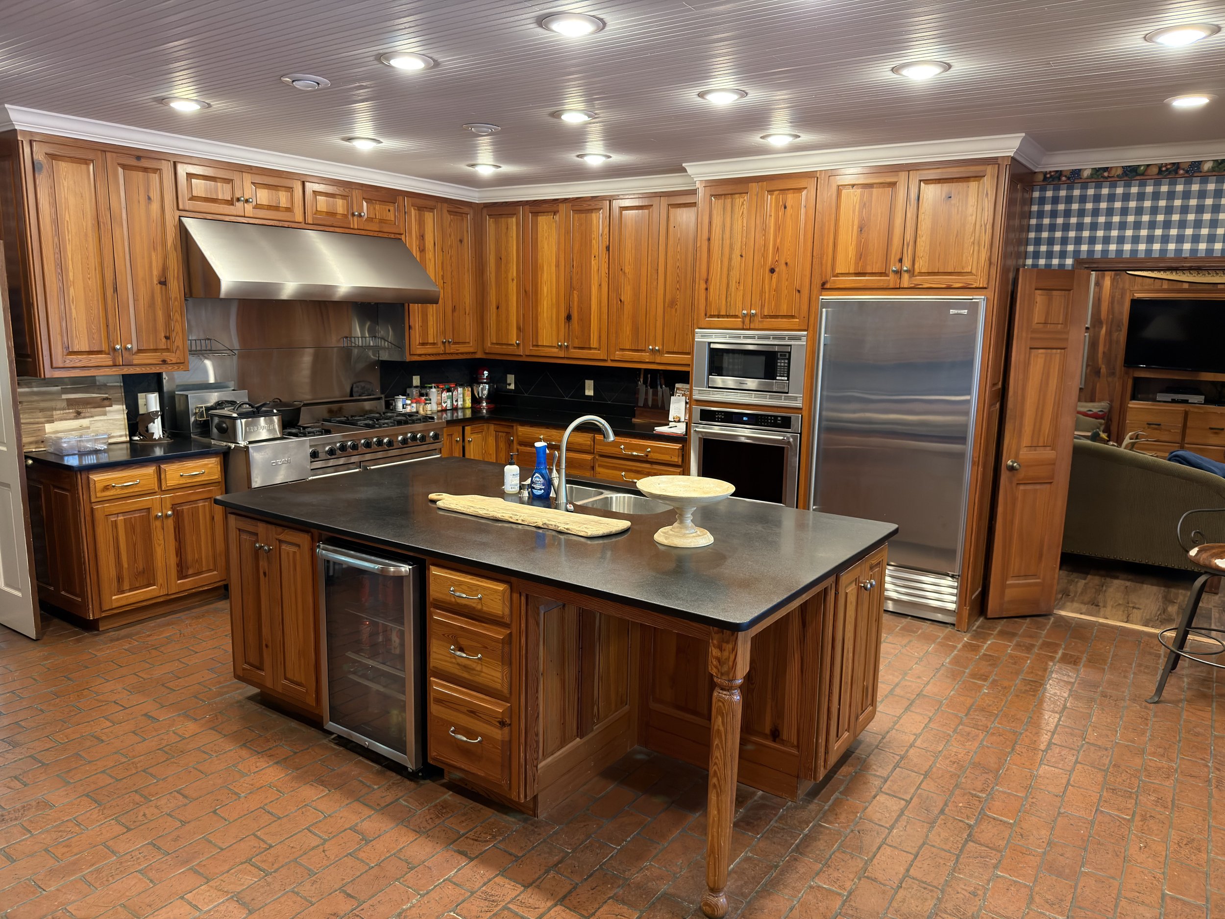 A spacious kitchen with wooden cabinets, a large kitchen island with a black countertop, stainless steel appliances, and brick flooring. Recessed lighting is installed in the ceiling.