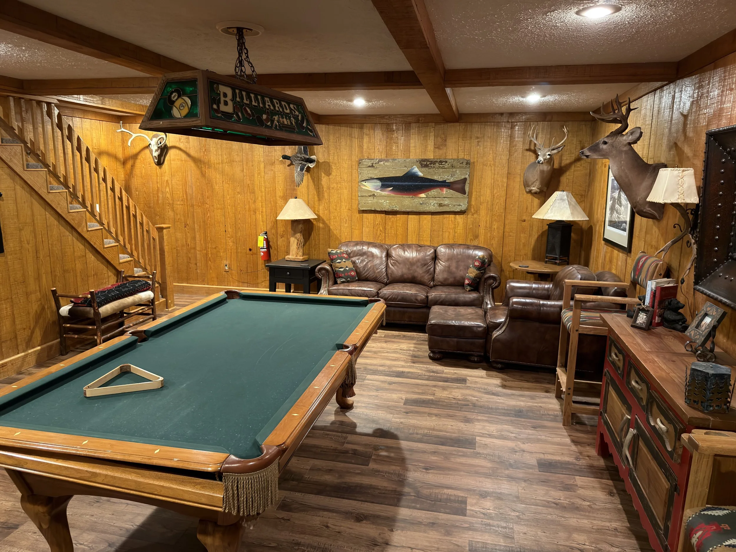 A recreational room with wood-paneled walls and a wood ceiling, featuring mounted deer and an oryx skull, a large colorful fish painting, leather chairs, and a pool table in the foreground.