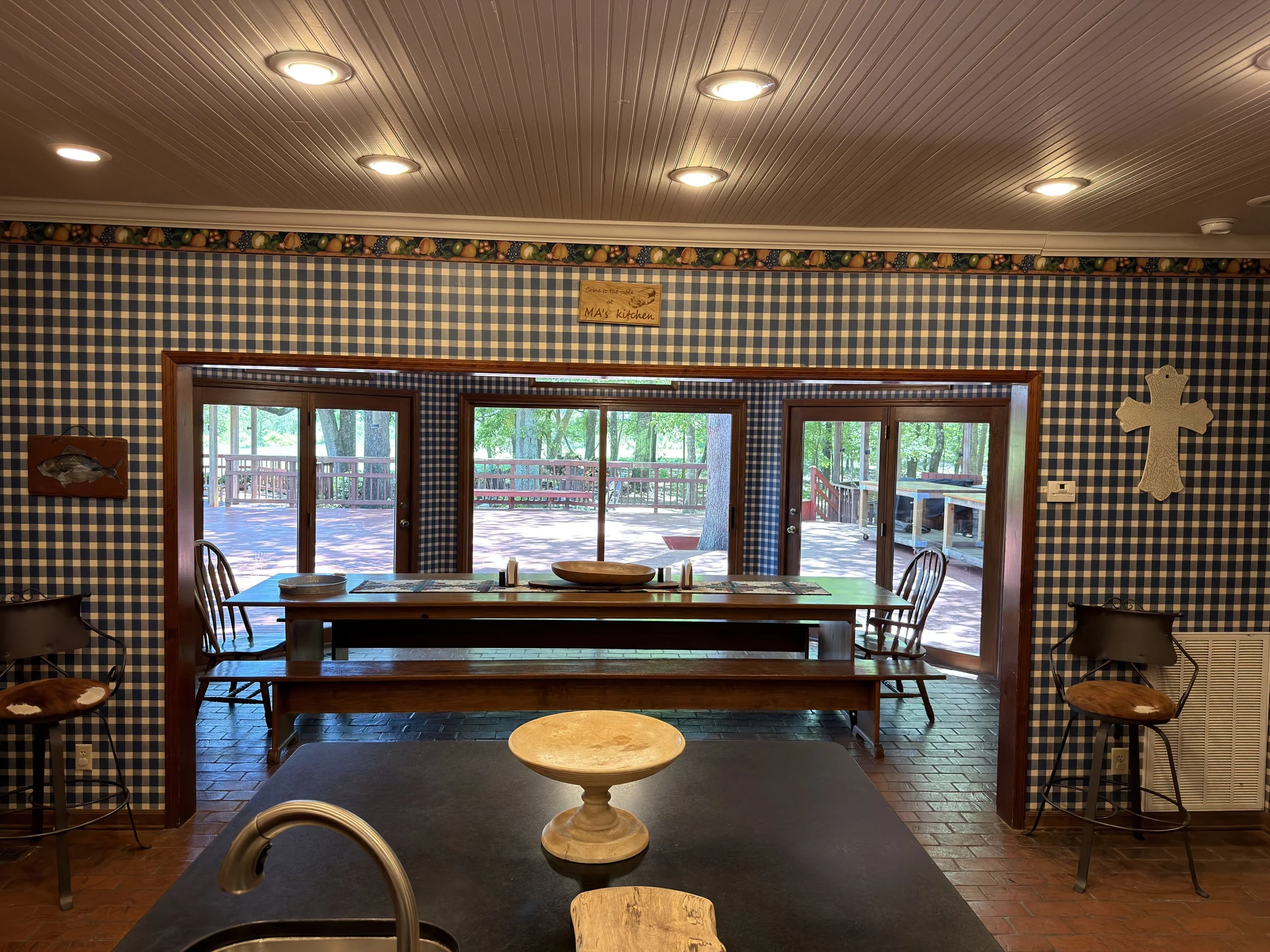 The image shows the interior of a room with a vaulted ceiling with lighting, blue and white checkered wallpaper, a long wooden dining table with chairs, and sliding glass doors opening to a deck with trees outside.