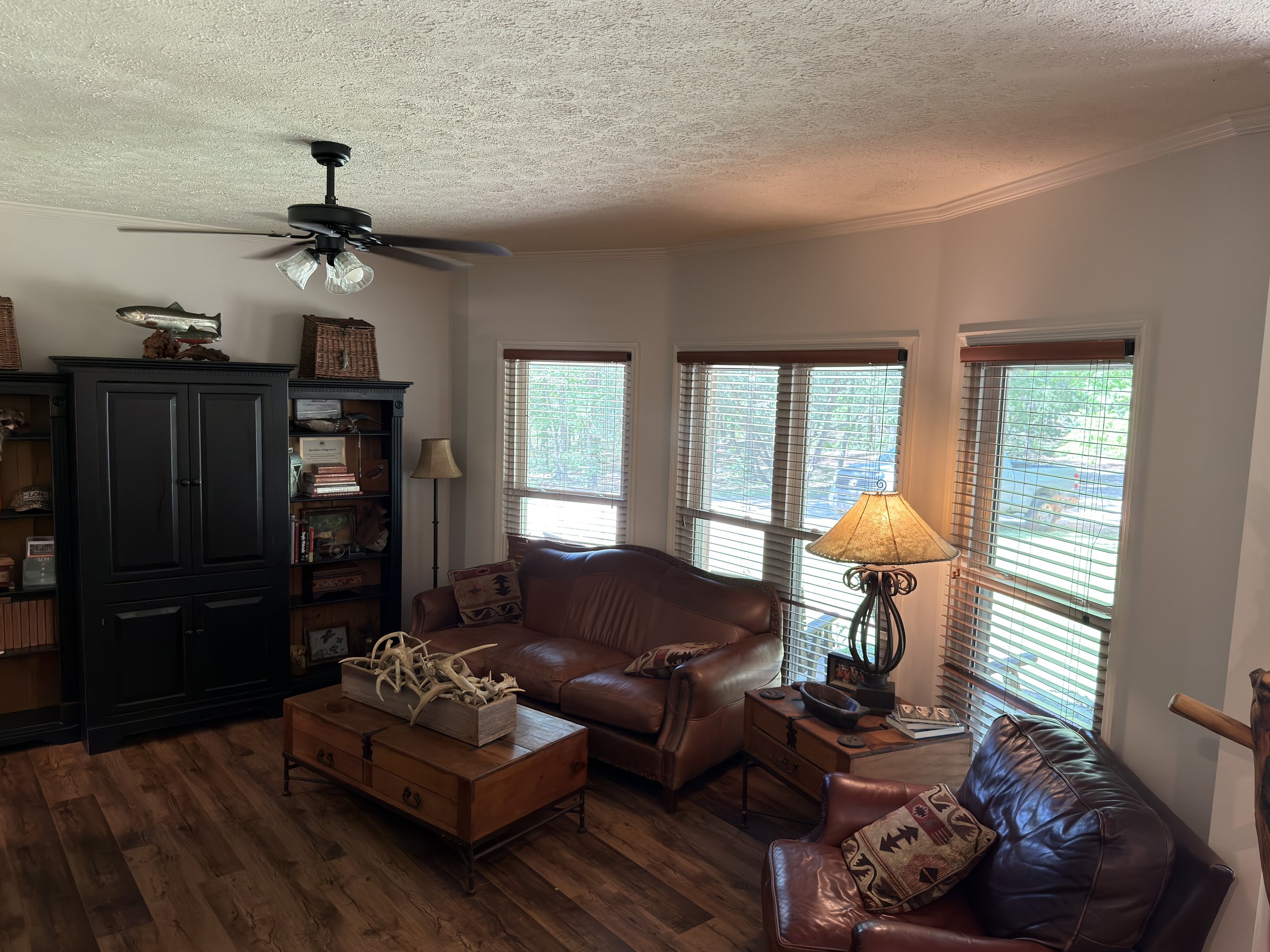 Living room with large windows, a leather couch, a wooden coffee table with decorative bones, and a tall dark bookshelf with navigational books and decorative items. Overhead ceiling fan with lights, and lamp on side table. Wooden flooring.