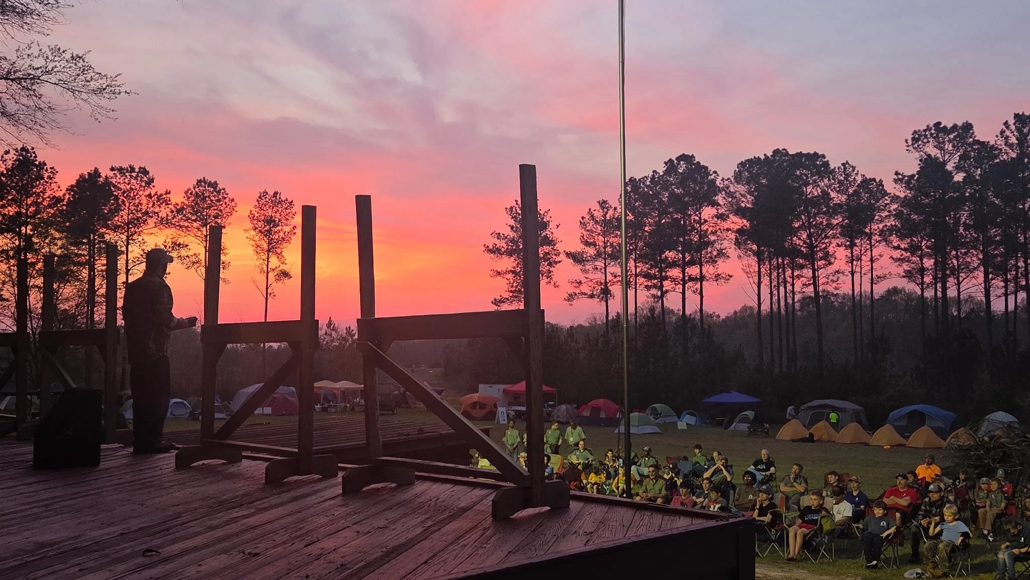 People gathered outdoors in tents during sunset, with a stage and a person speaking in front of a seated audience, surrounded by trees.