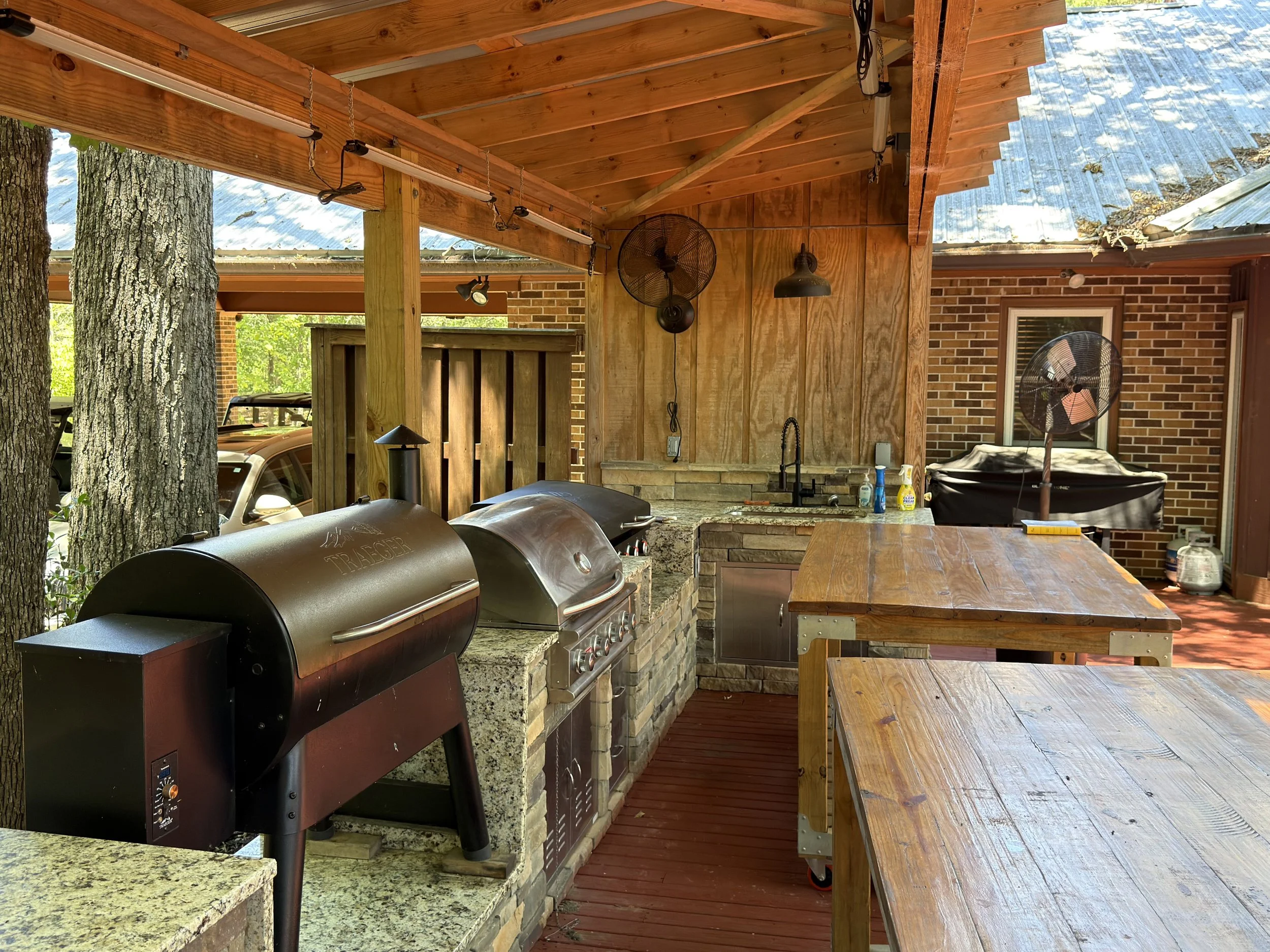 An outdoor kitchen area with a wooden roof, brick walls, a granite countertop, and a barbecue grill, surrounded by trees and a wooden table.