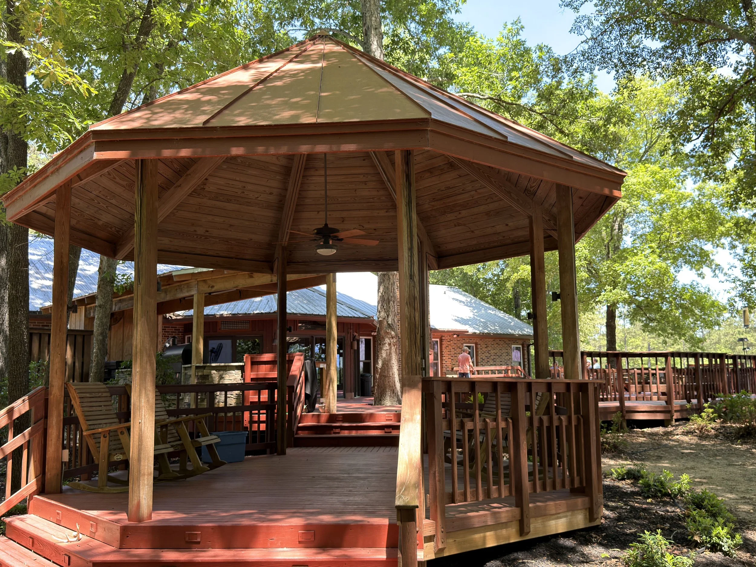 A wooden gazebo with a tan roof and ceiling fan, surrounded by trees in a backyard with a deck and house in the background.