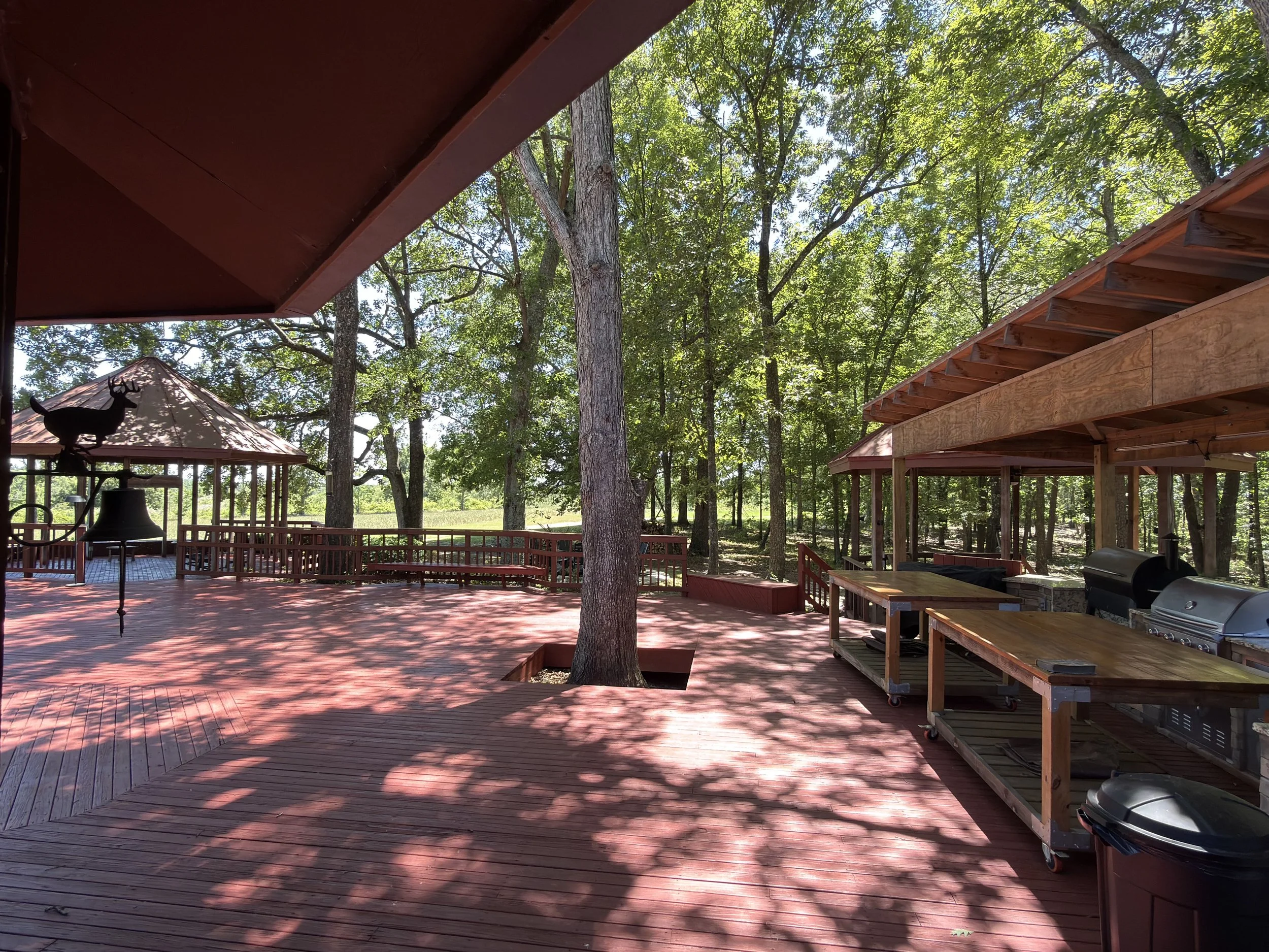 A wooden deck in a backyard with trees, a metal deer sculpture on a roofed structure, and outdoor furniture and grilling equipment. Shadows from trees are on the deck.