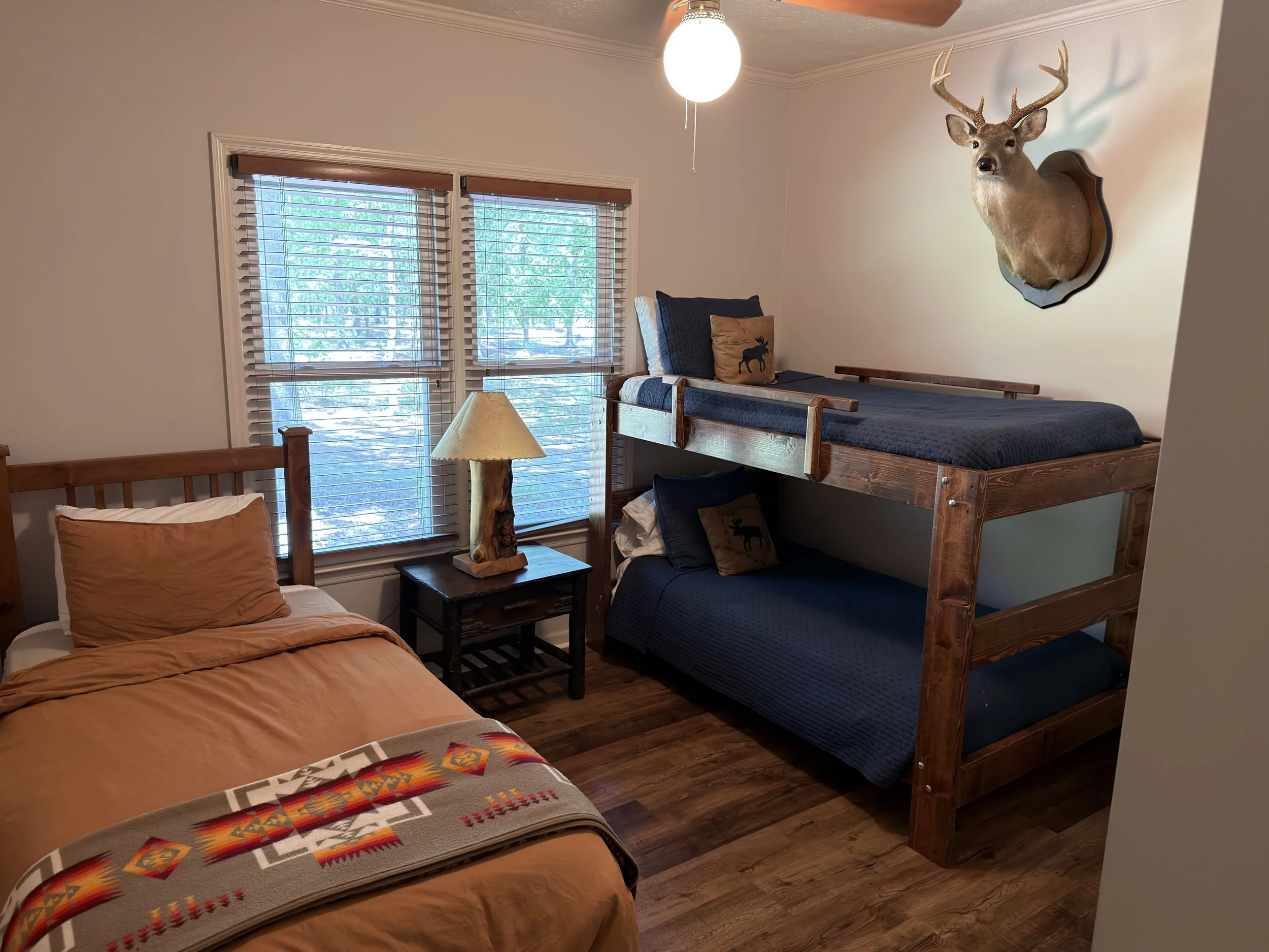 A bedroom with a bed in the foreground covered with a tan bedspread and a colorful patterned blanket at the foot. To the right, a bunk bed made of wood with dark blue bedding and pillows featuring moose images. A small wooden nightstand with a rustic