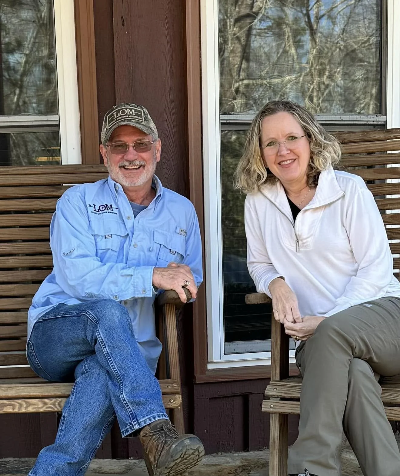 A smiling older man wearing a baseball cap, glasses, and a blue button-up shirt, sitting on a wooden bench with a woman with curly blonde hair and glasses, wearing a white jacket and khaki pants, outdoors on a wooden deck with a wooded background.