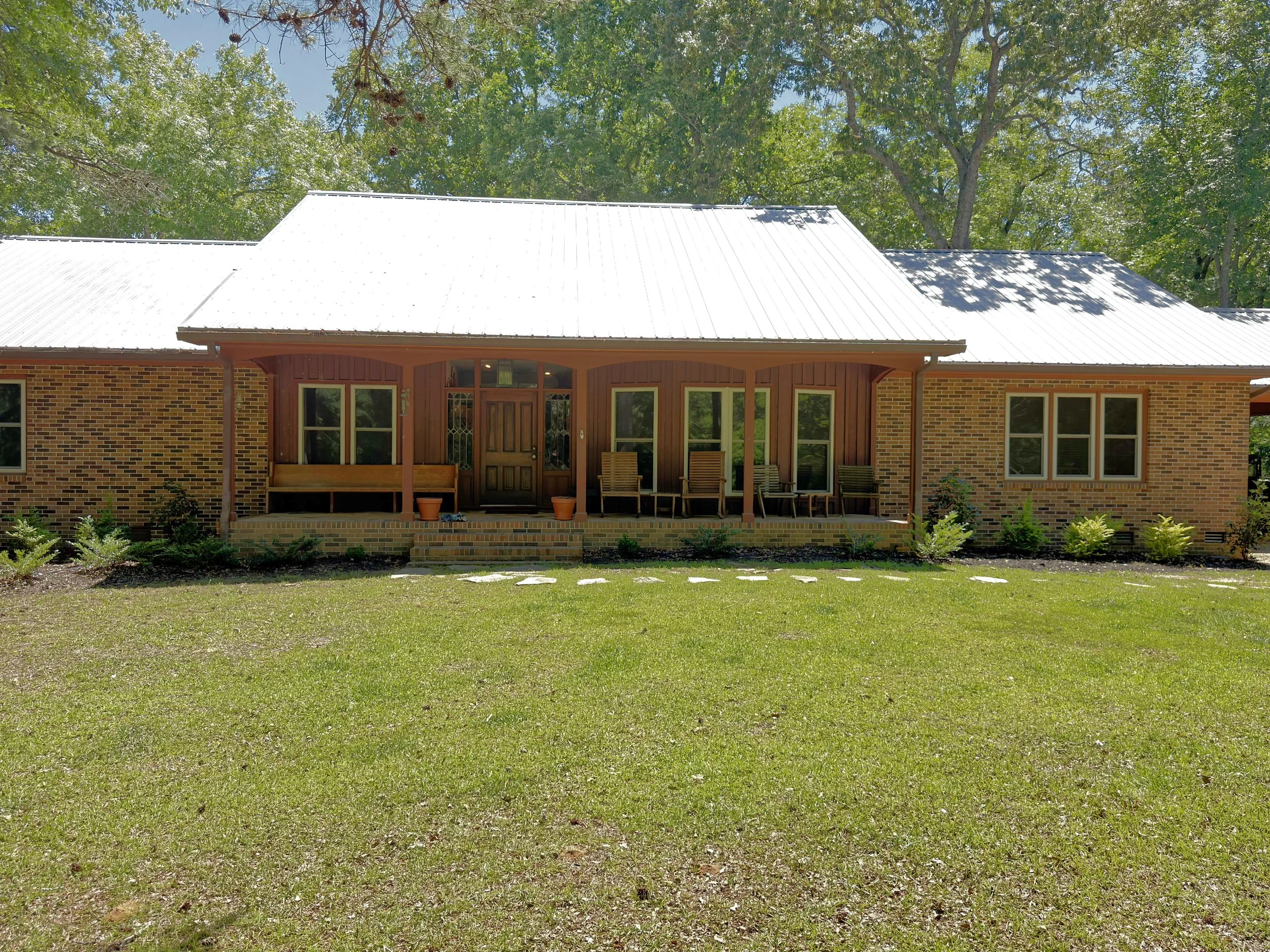 Front view of a house with a metal roof, brick walls, a wooden front door, and large windows, surrounded by a green lawn and tall trees in the background.