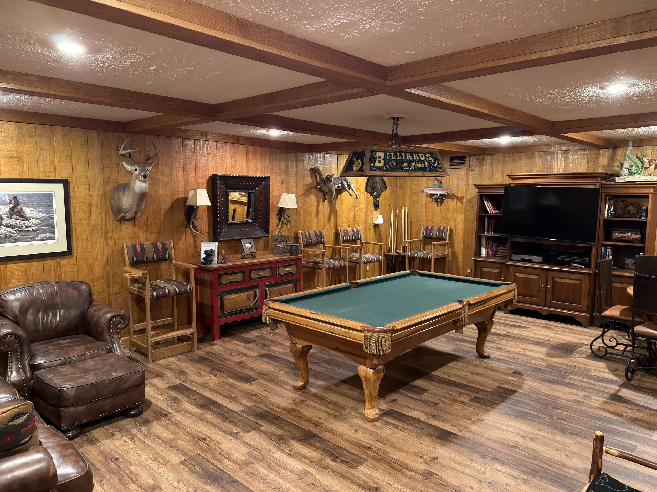 A cozy basement room decorated with hunting-themed items including mounted deer and bears, fishing decor, and wooden trophies. There is a pool table, a leather armchair, multiple chairs with patterned upholstery, a TV and bookshelf, and warm wooden p