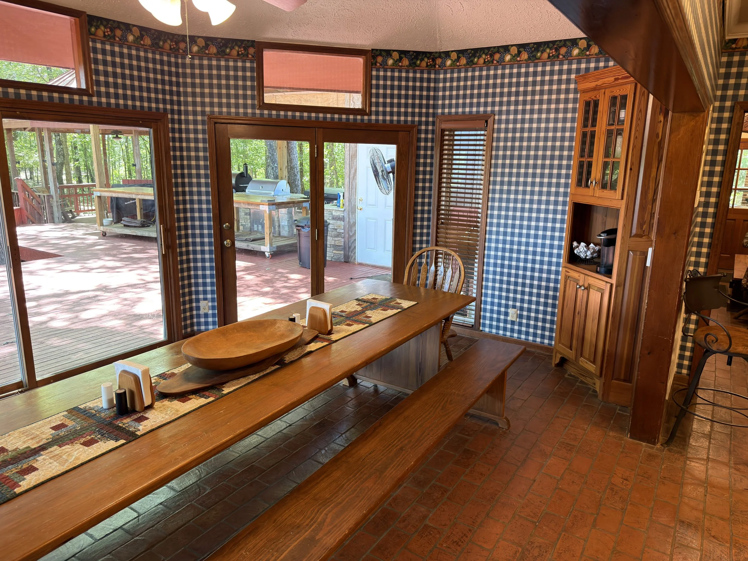 Kitchen area with checkered blue and white wallpaper, wooden cabinet, glass door leading to outdoor deck with grill, wooden dining table, and windows overlooking a wooded backyard.
