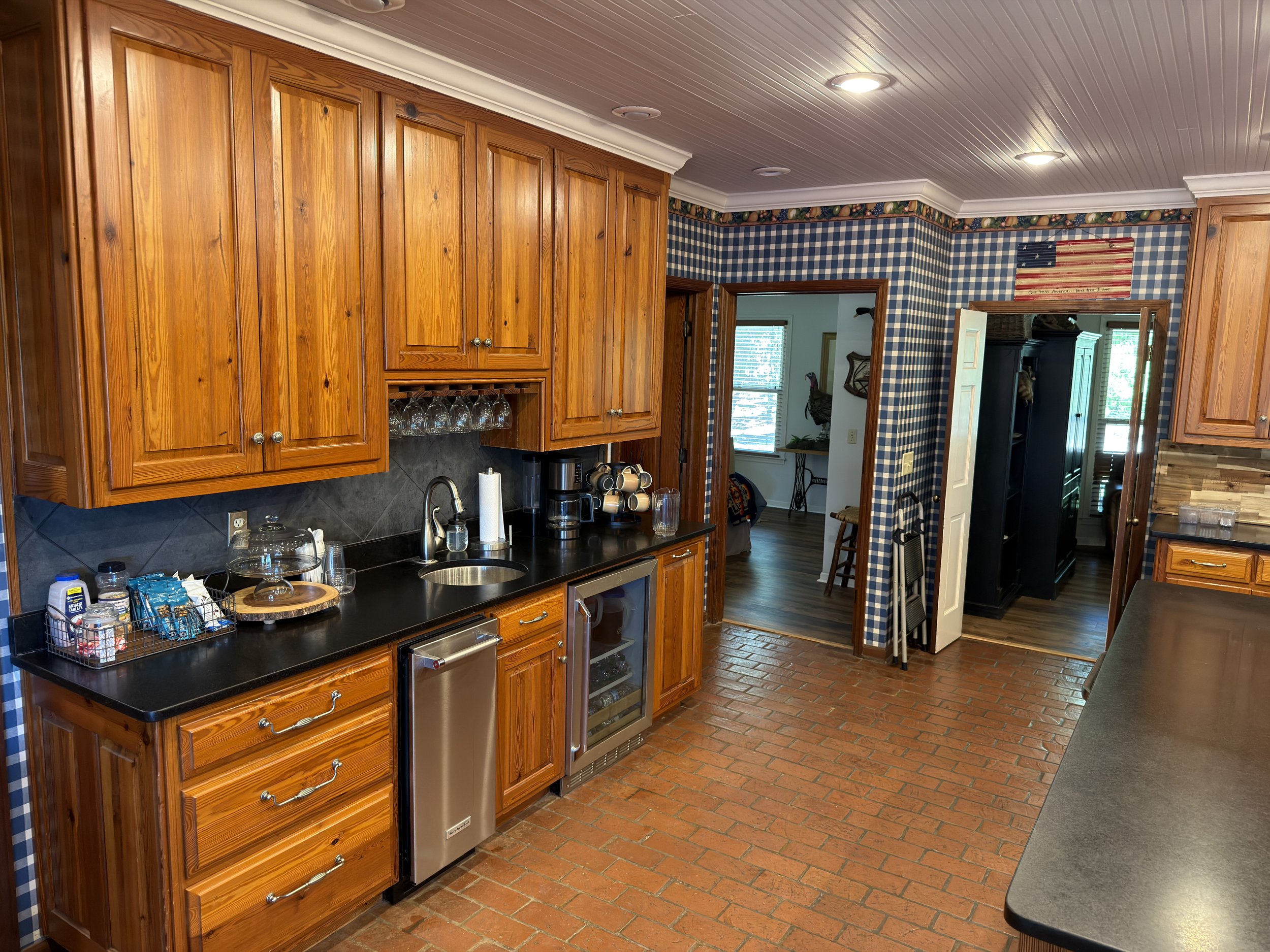 Kitchen with wooden cabinets, black countertop, and brick floor, featuring a coffee maker, glassware, and a partially visible open door leading to another room with a window.