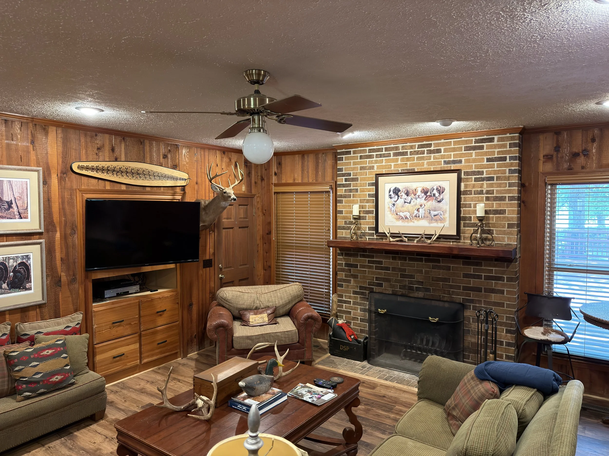 A cozy living room with wood-paneled walls, a brick fireplace, and a ceiling fan with a light. The room features furniture including a leather armchair, a sofa with patterned pillows, and a wooden coffee table. Decorations include framed pictures, a 