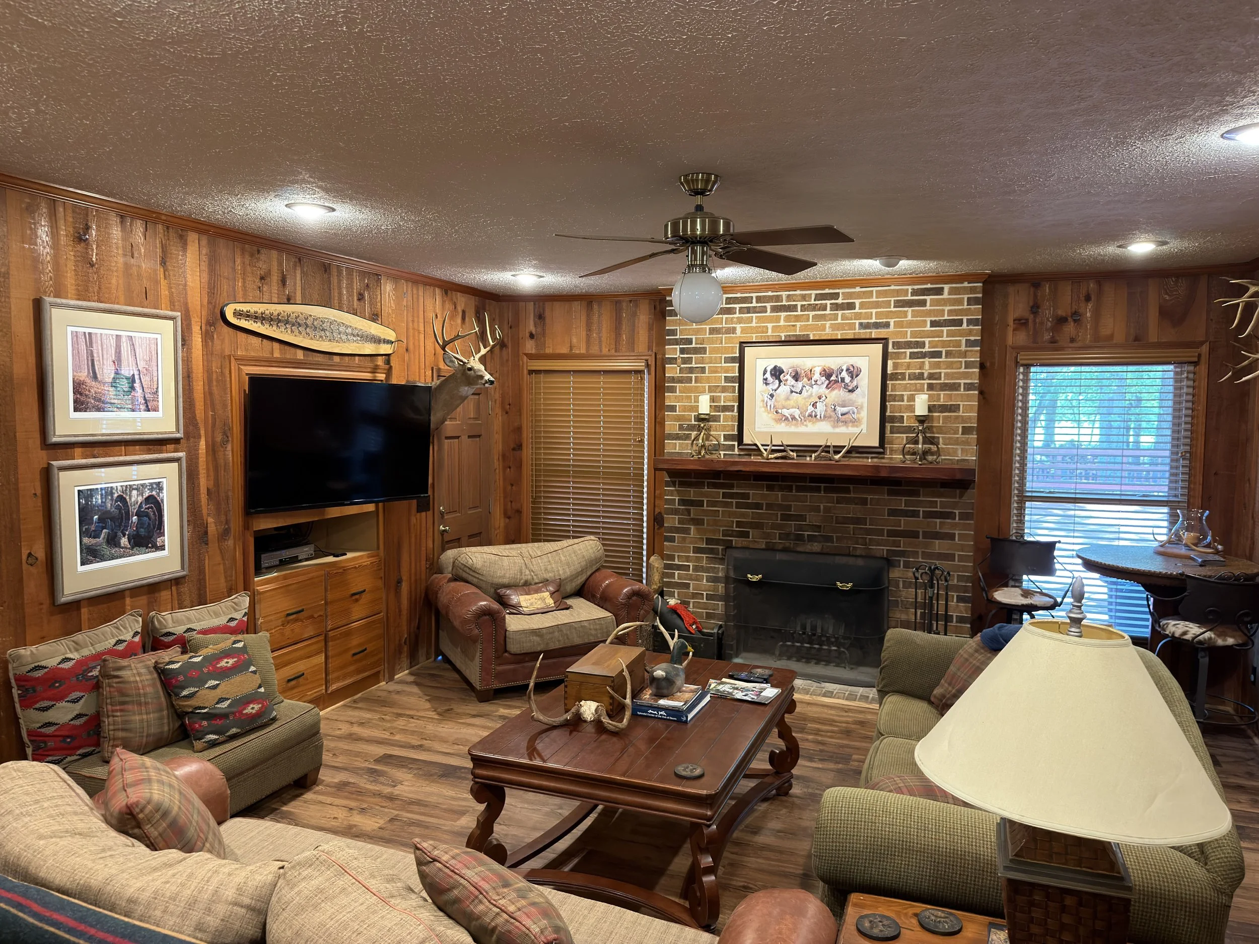 A cozy living room with wooden panel walls and a brick fireplace, decorated with wildlife-themed art and antlers. There are multiple sofas and chairs with plaid cushions, a wooden coffee table with books and decorative items, and a television mounted