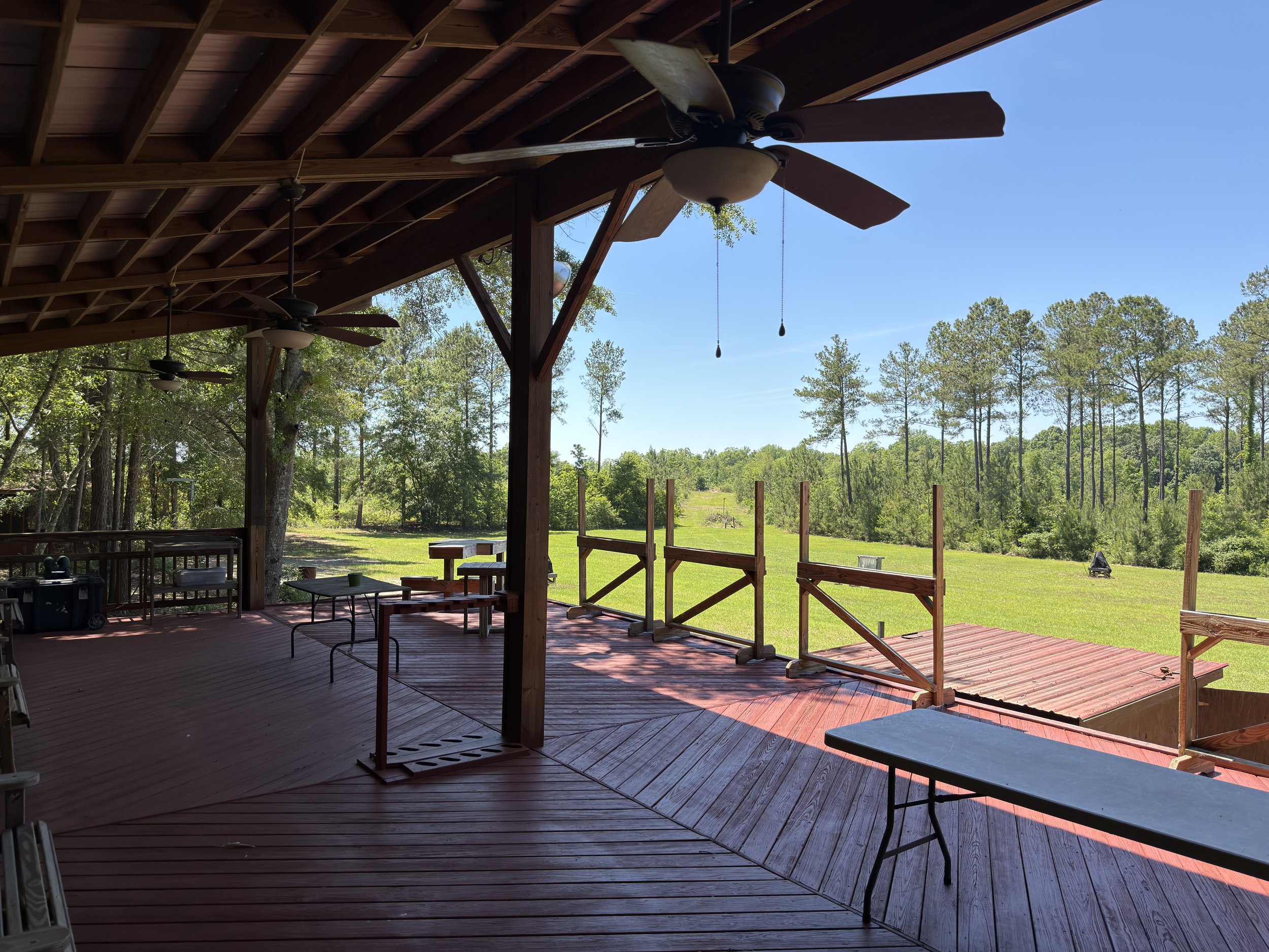 A wooden porch with ceiling fans, overlooking a green backyard with trees and a blue sky.