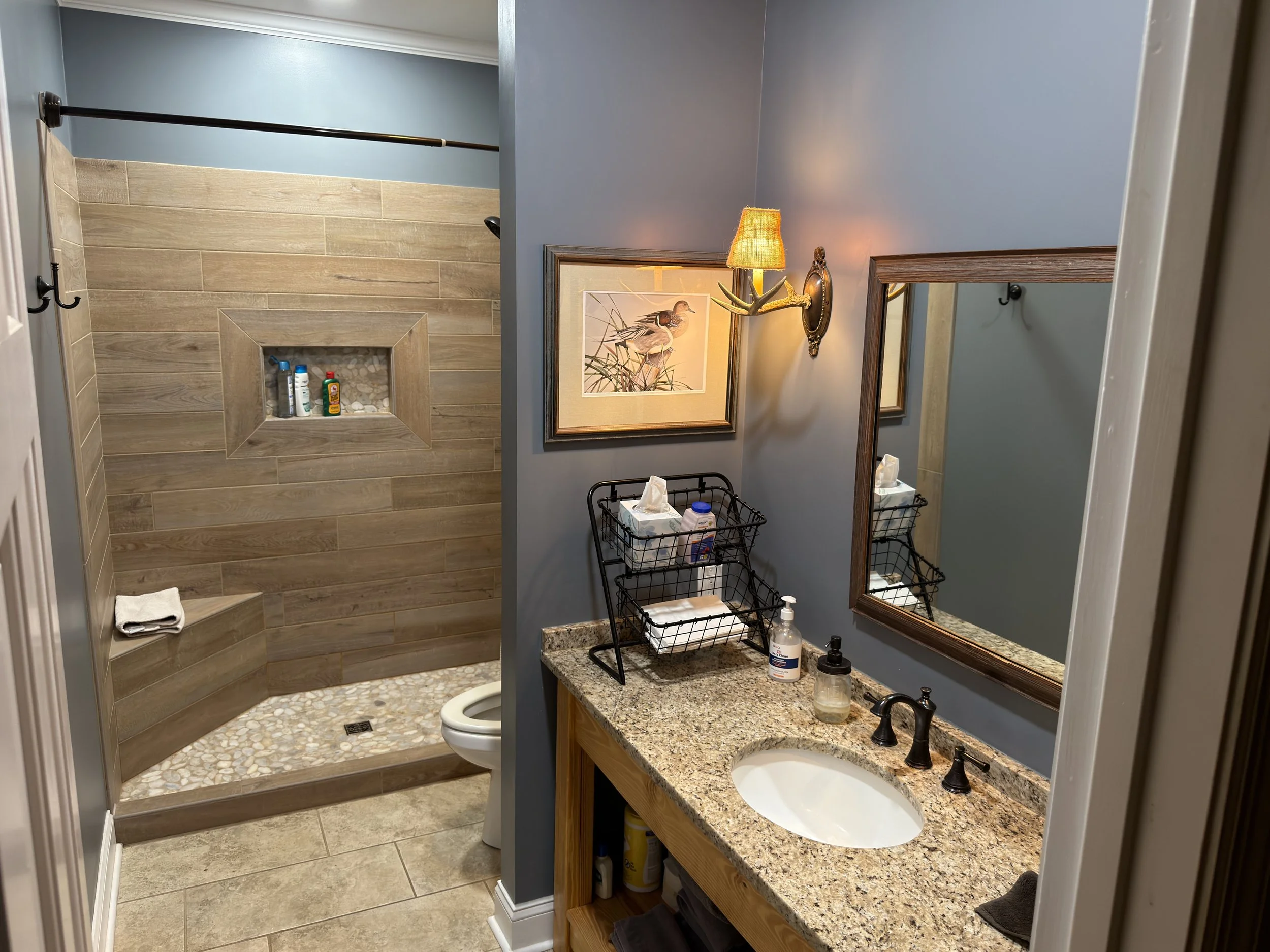 Bathroom with a walk-in shower on the left, a granite countertop with an undermount sink and a framed mirror above it on the right. There are two wicker wall-mounted light fixtures, a framed artwork of a bird on the wall, and two wire baskets on the 
