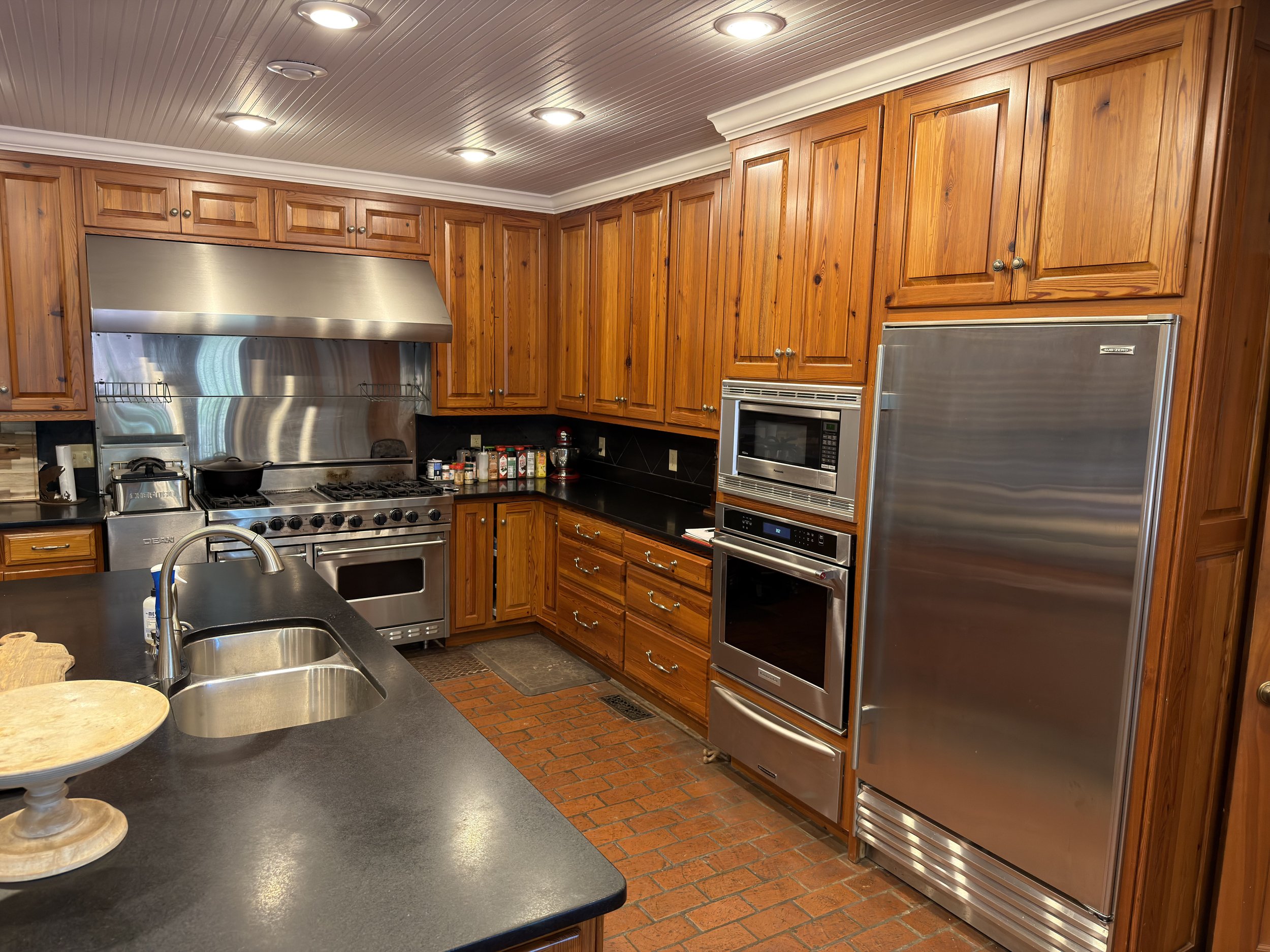Kitchen with cherry wood cabinets, stainless steel appliances including a stove, microwave, and refrigerator. Recessed ceiling lights, black countertop, and a brick floor.