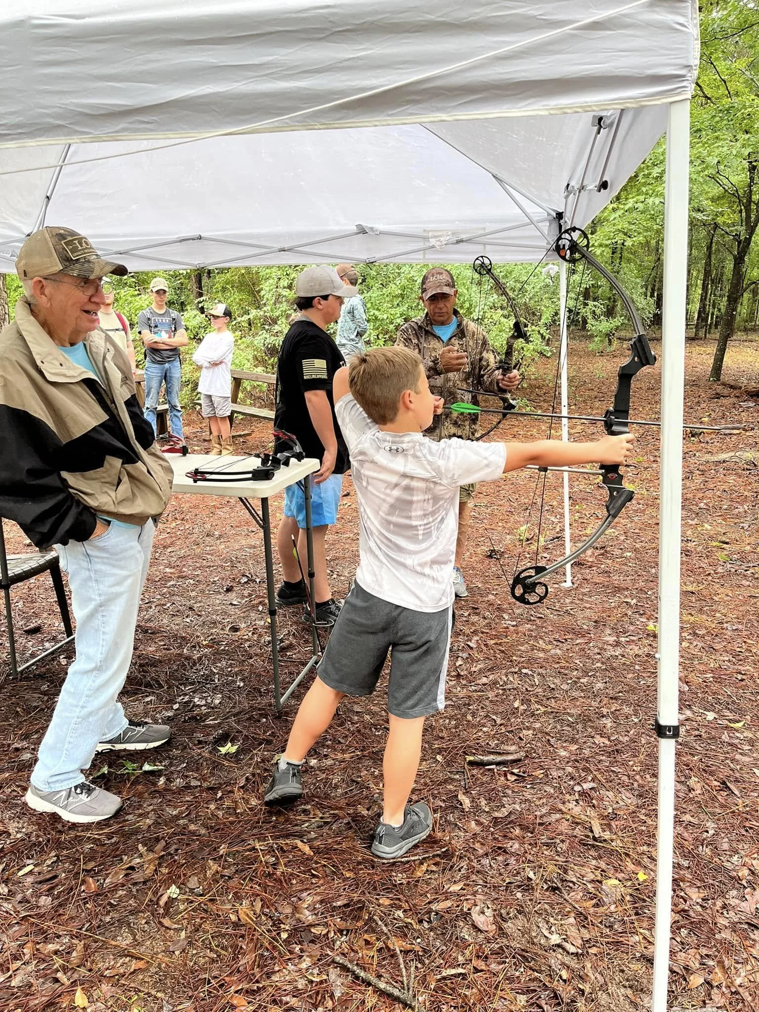 A group of people in an outdoor wooded area participating in archery practice under a white canopy. A young boy is drawing a bow, aiming at a target, while adults observe, some smiling and others watching attentively.