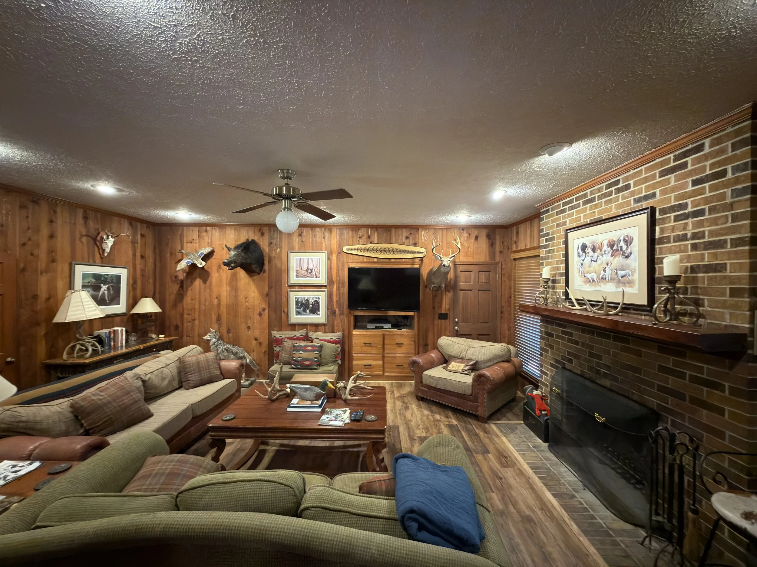 Living room with wood-paneled walls, hunting decor including animal heads, framed pictures, and a large flat-screen TV. There are sofas and armchairs with plaid and solid cushions, a coffee table with magazines, and a brick fireplace with decorative 