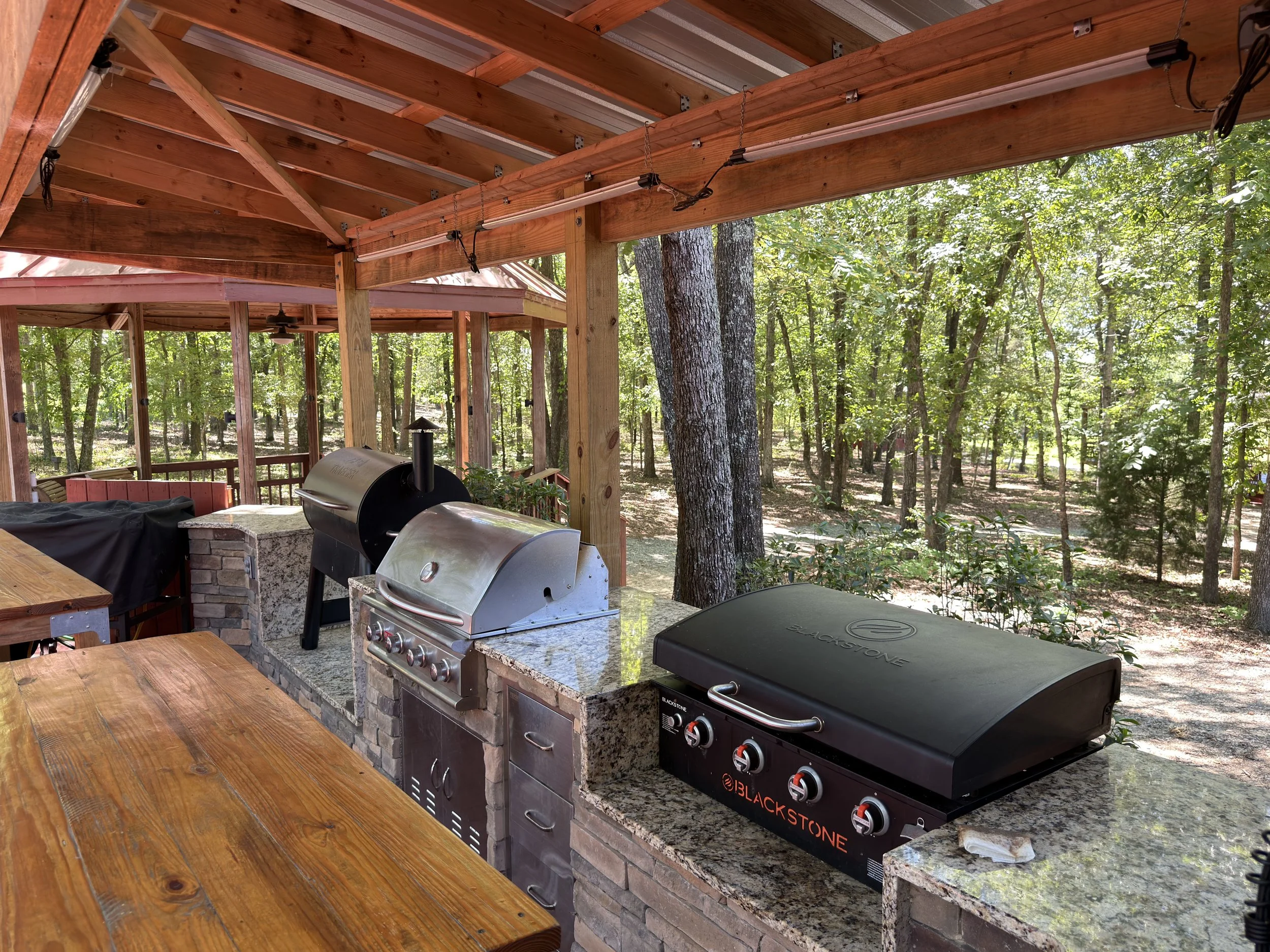 Outdoor wooden deck with a propane grill and a Blackstone griddle, surrounded by trees and dense forest, with a wooden roof overhead.