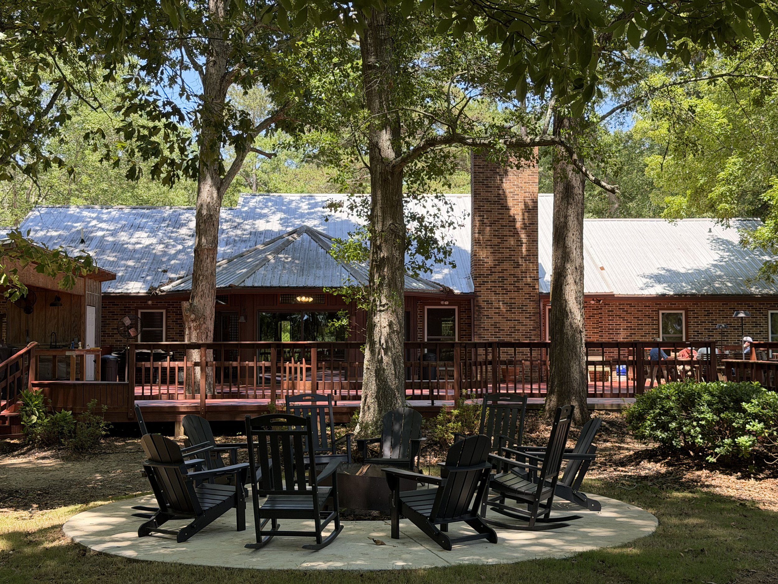 A backyard patio with black Adirondack chairs surrounding a fire pit, with trees providing shade and a house with a brick chimney and a metal roof in the background.