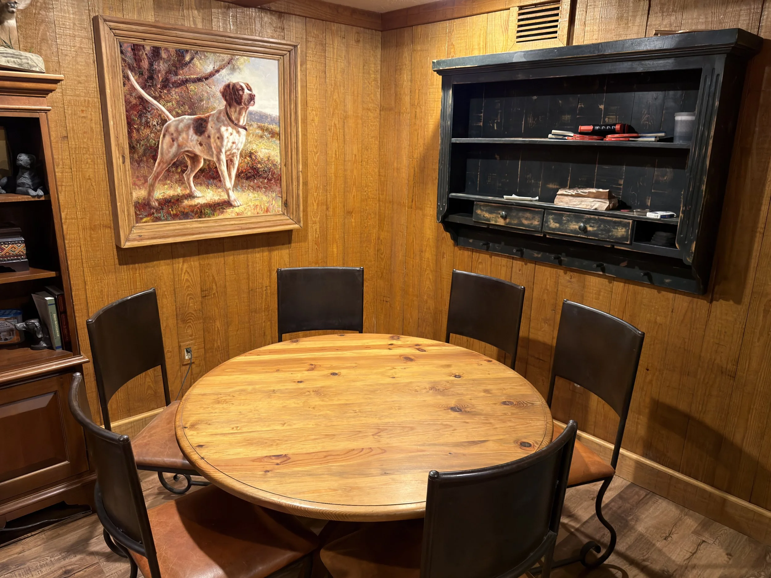 A wooden dining table with five black chairs around it, wood-paneled walls, a large dog painting, and a black wooden wall shelf with books and items.