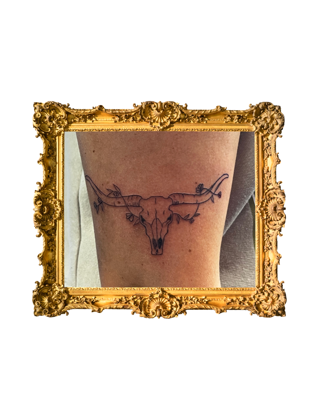 Tattoo of a longhorn steer skull with floral and vine details on a person's upper arm, framed with a gold ornate picture frame.