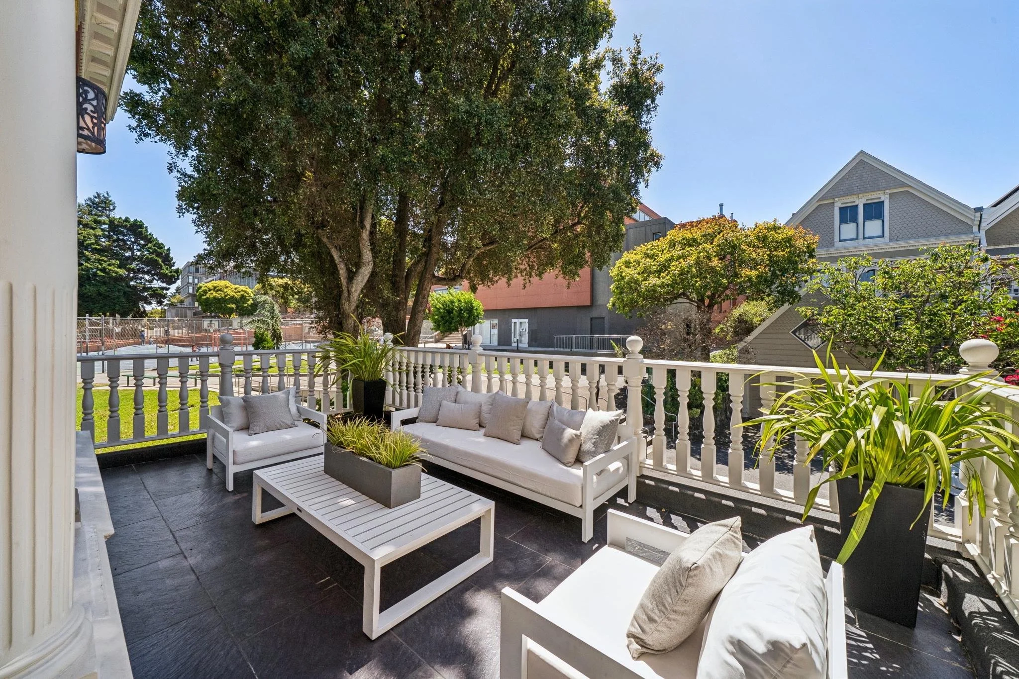 A balcony with outdoor seating, including white sofas with beige cushions, a coffee table, potted plants, a white railing, and a view of large trees and neighboring houses under a clear blue sky.