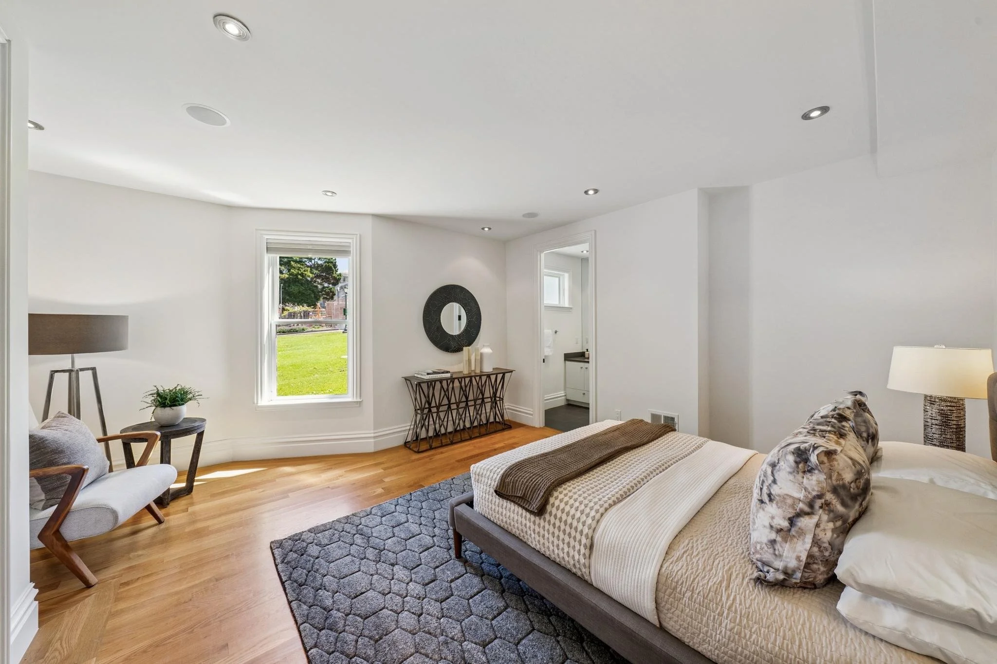 Living room with white walls and large windows showing a grassy yard outside. Furniture includes a sofa with patterned pillows, a side chair, a small table, and a coffee table. Decorated with a mirror, table lamp, and vases. Hardwood floors and ceili
