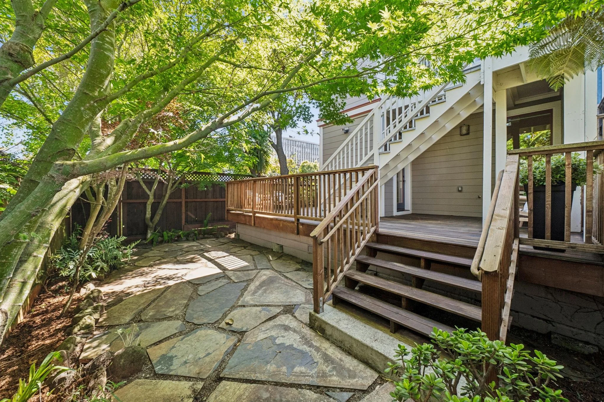 A backyard area with a stone-paved ground, a wooden deck with stairs, and lush green trees and plants providing shade.