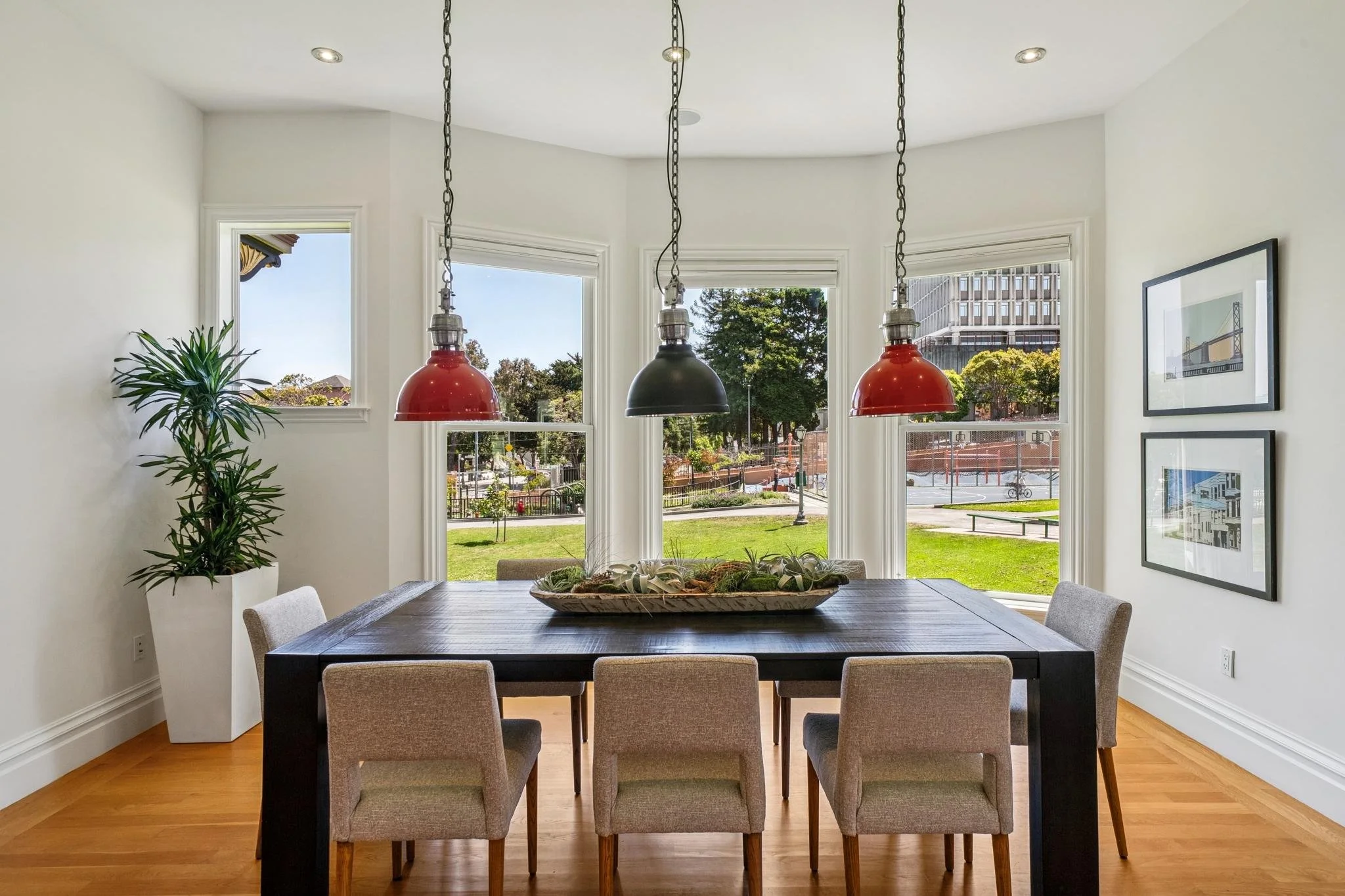 A modern dining room with a dark wood table, beige chairs, and three hanging pendant lights (red, black, red). Large windows with white frames reveal a view of a park with trees and a basketball court. There is a tall green plant in a white pot on th