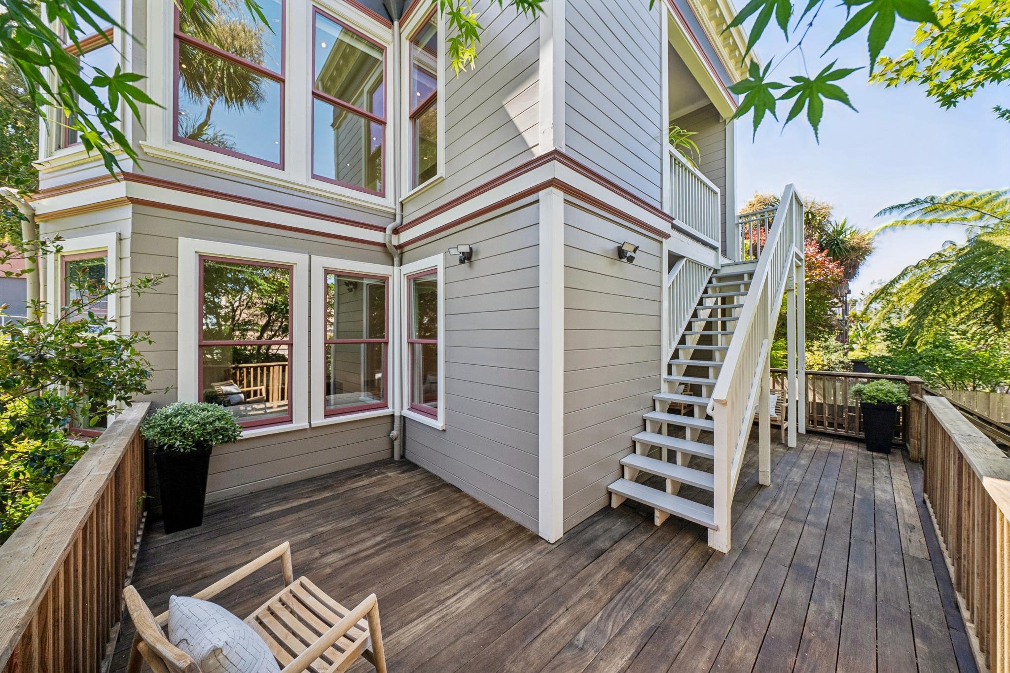 Outdoor wooden deck with seating, planter, and stairs leading to a house with beige siding and multiple windows, surrounded by green trees.