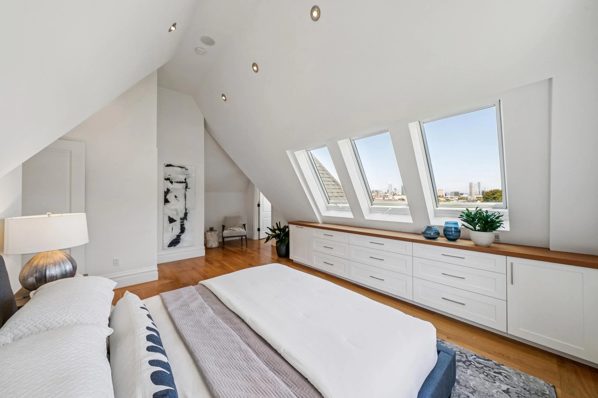 A bright white bedroom with sloped ceilings and three large skylight windows overlooking a cityscape. The room has a white built-in dresser with plants and decorative items, and a bed with white bedding and navy blue accents.
