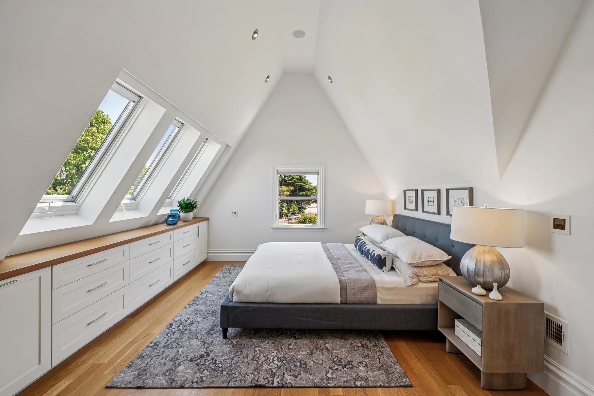A bedroom with a sloped ceiling, white walls, three windows on the left, and a single window at the back showing greenery outside. The bed has a dark upholstered headboard, white bedding, and pillows. A wooden nightstand on the right has a lamp and d