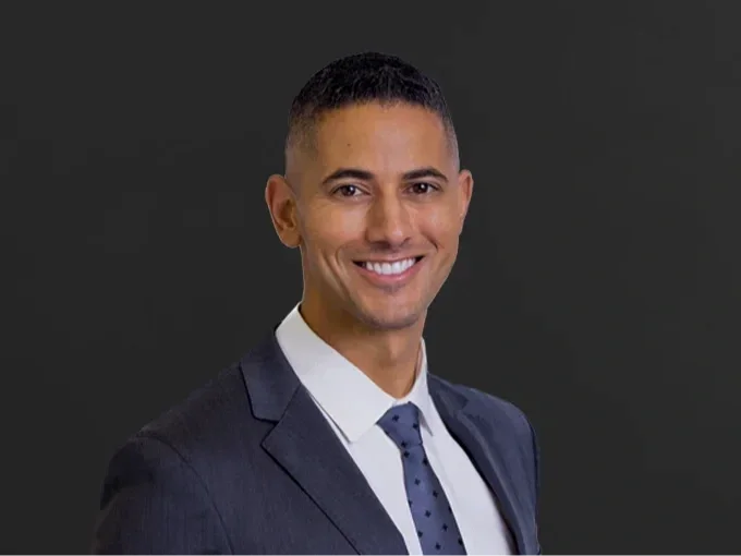 Professional headshot of a smiling man in a suit and tie against a dark background.