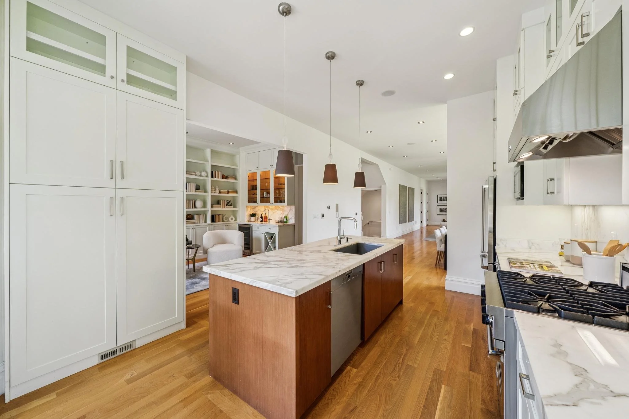 Open concept kitchen with white cabinetry and wood flooring, featuring a marble kitchen island with a stainless steel sink, modern black kitchen range, and pendant lighting, with a sitting area and built-in open shelves in the background.