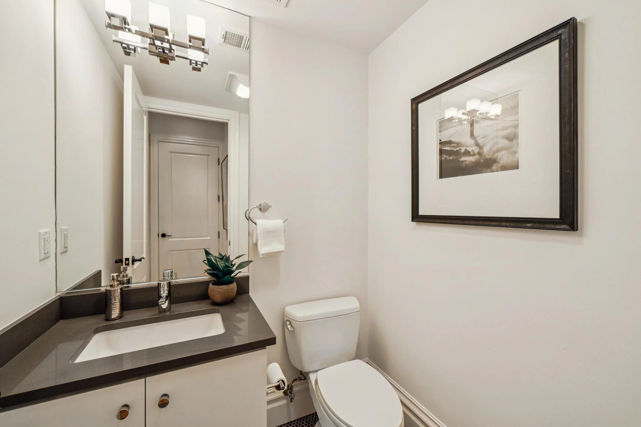 Small bathroom with white walls, a large mirror, a dark gray countertop with a sink, a small potted plant, soap dispensers, a towel on a chrome holder, a framed black and white photo of a bridge, and a white toilet.
