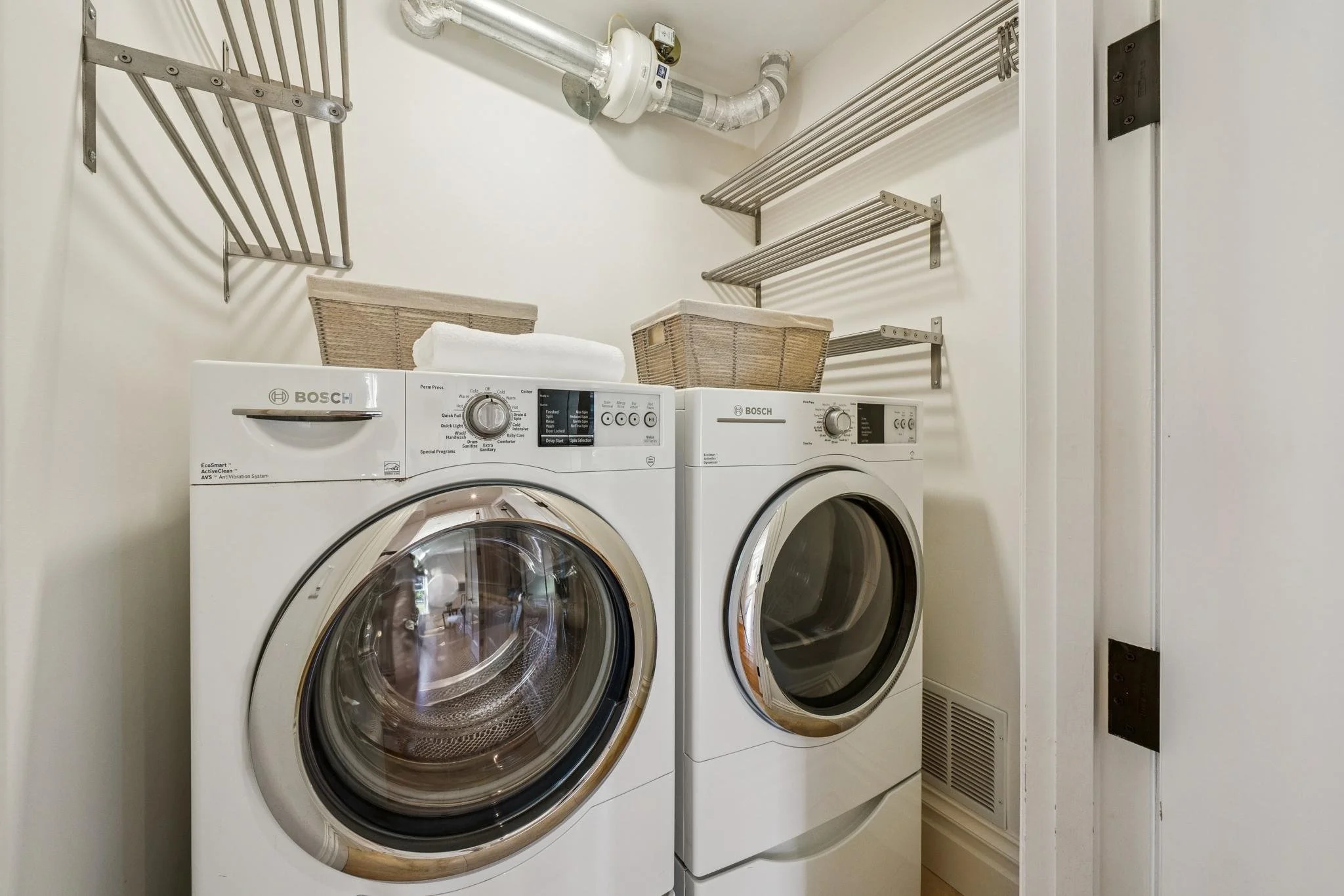 A laundry room with two front-loading Bosch washing machines side by side, with laundry baskets on top and metal shelving above, and a vent on the wall.