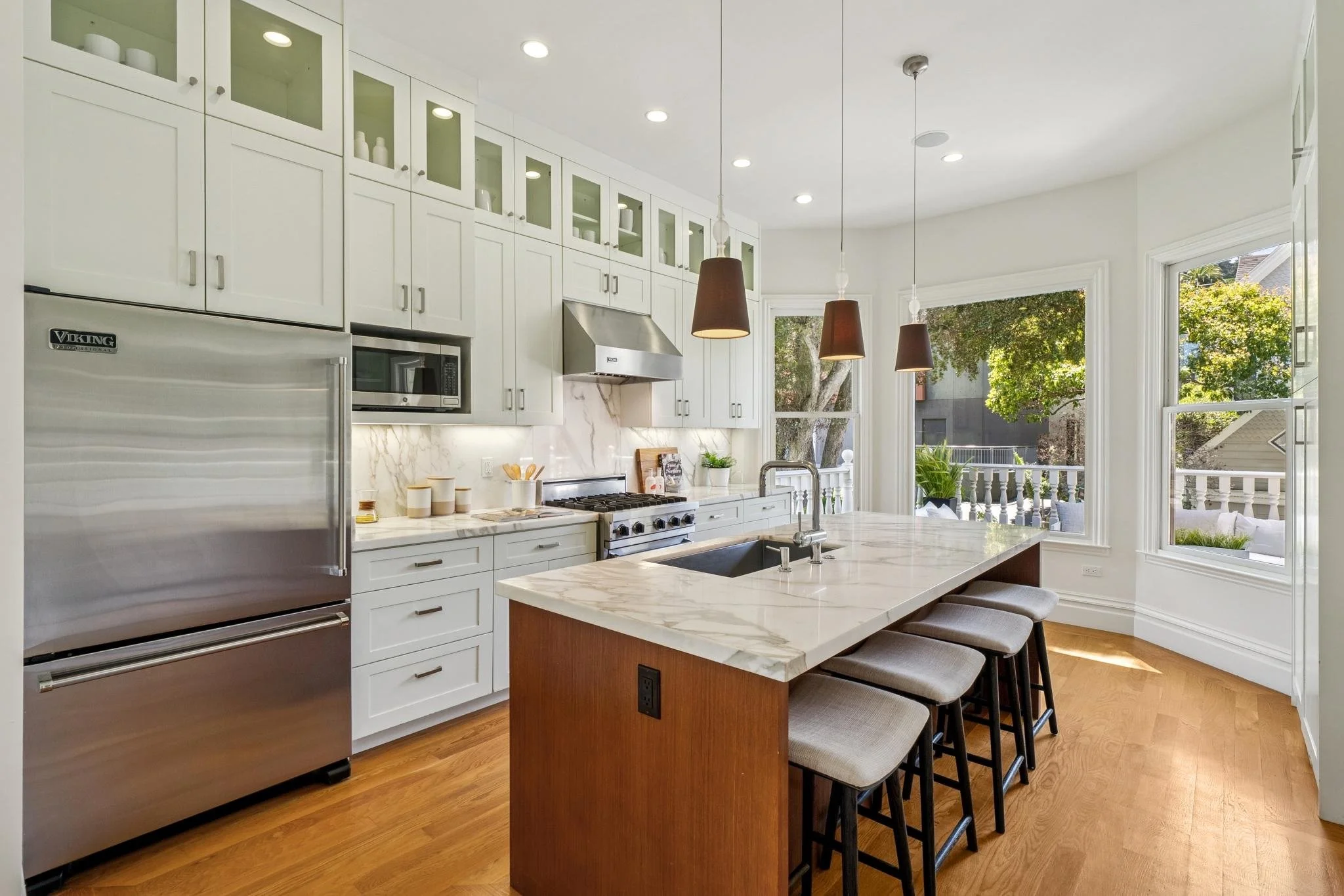 Modern kitchen with white cabinets, a marble countertop island, stainless steel appliances, and large windows showing trees outside.