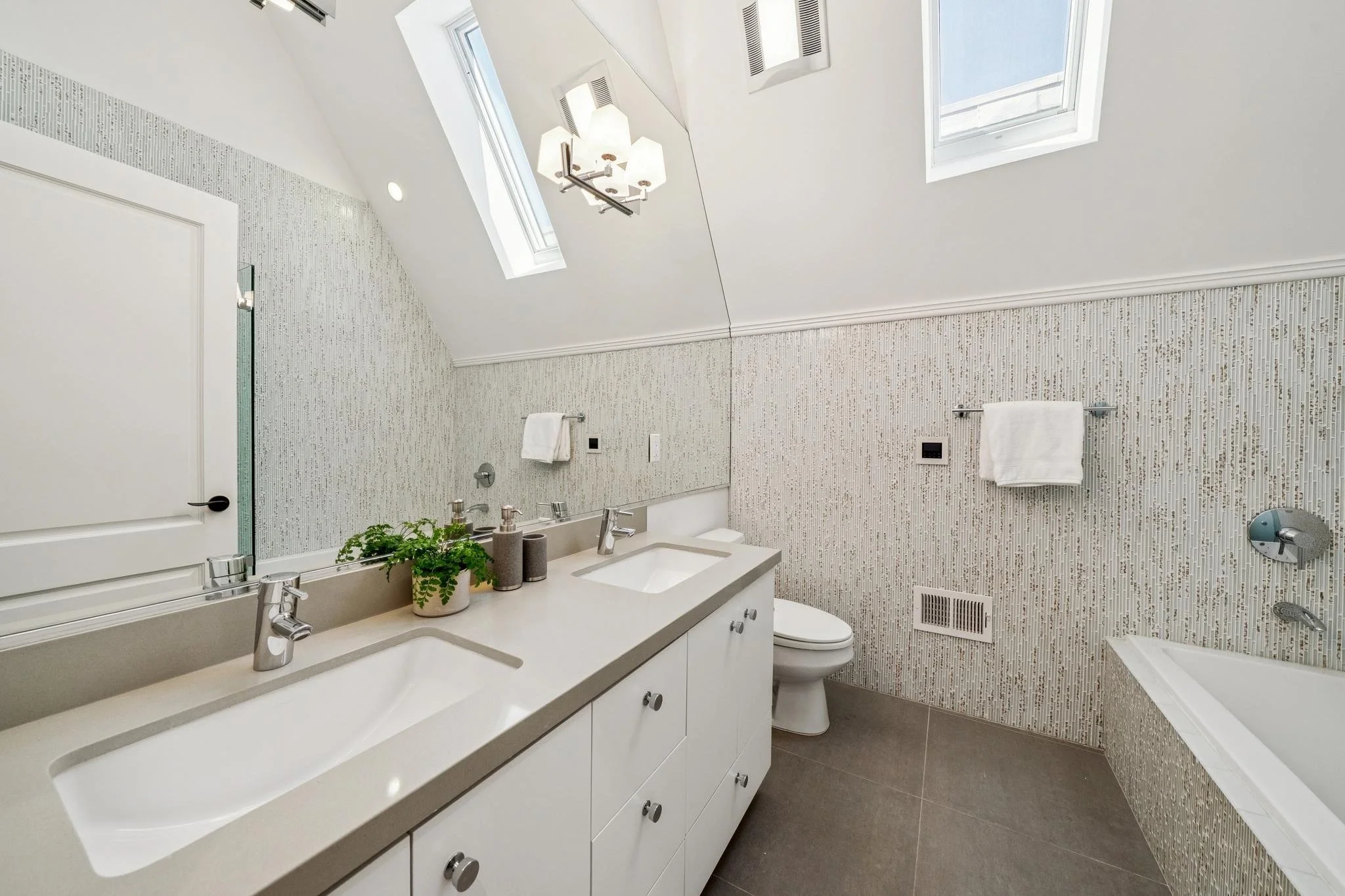 Bathroom with dual sinks, mirror, white cabinets, and mosaic tiled walls. Features skylight windows, multiple light fixtures, and a towel bar with a white towel.
