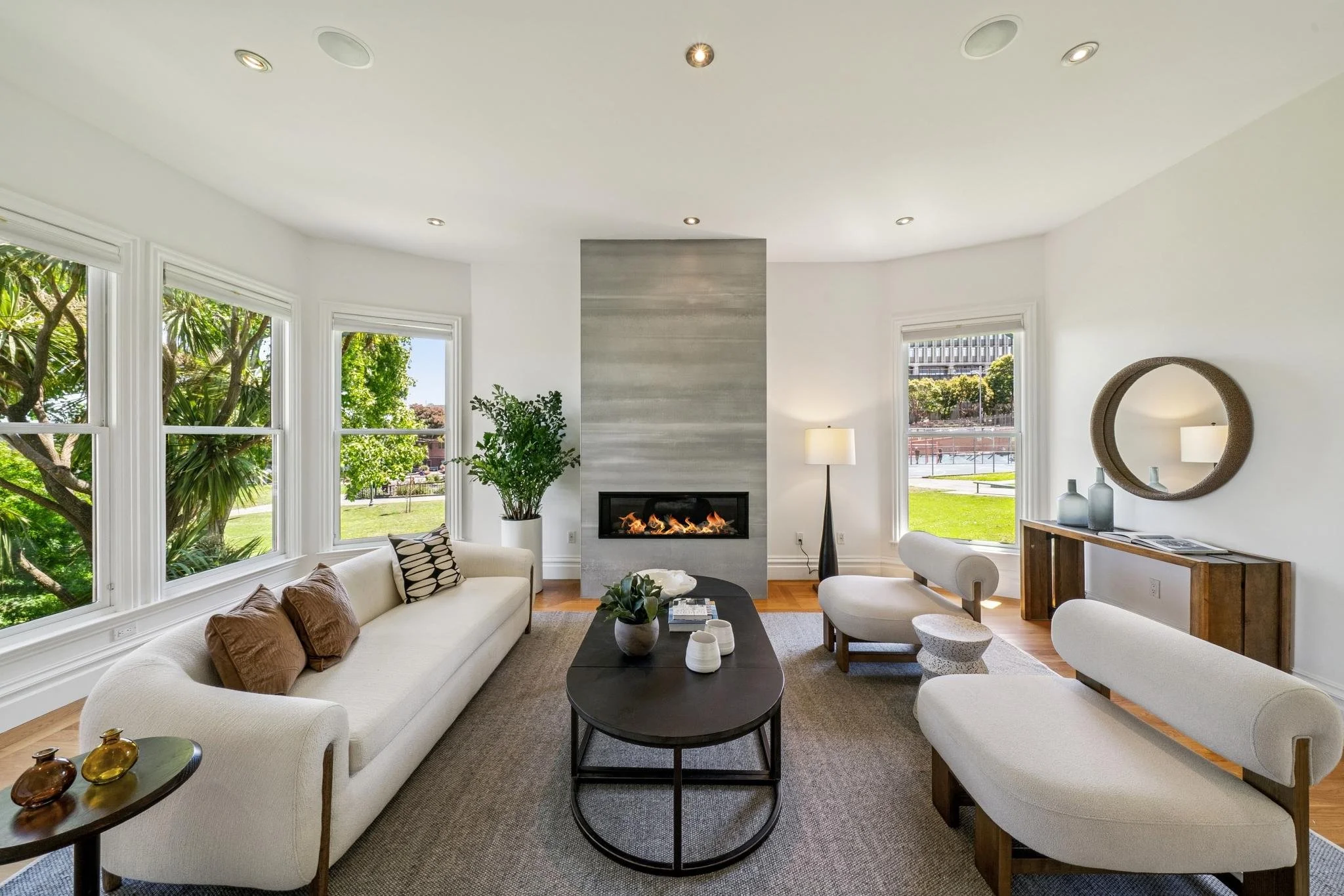 Modern living room with white walls and large windows showing green trees outside. Features include a fireplace, white sofas, a black coffee table, and a decorative round mirror on the wall.