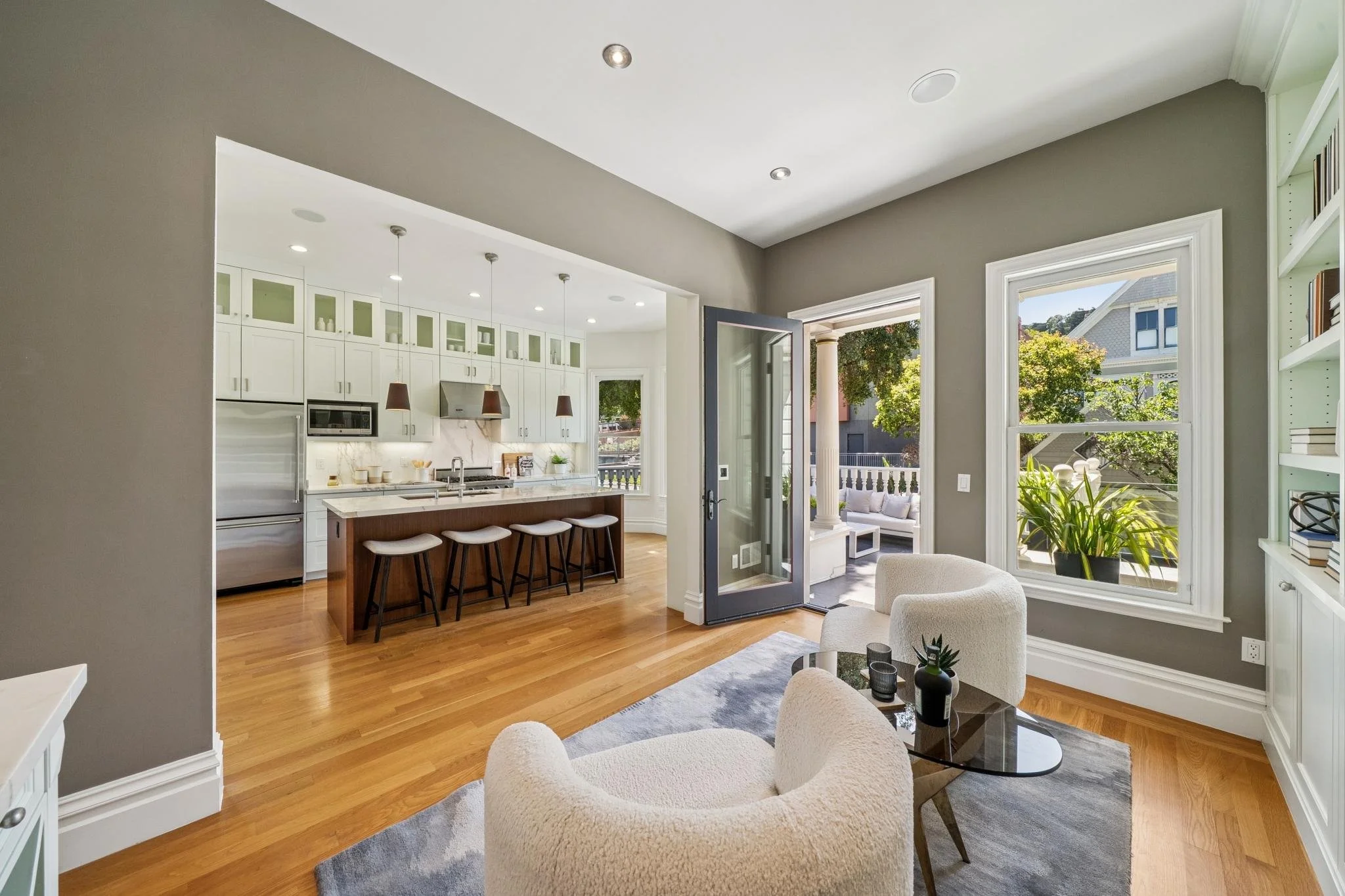 Open-concept living area with a view into the kitchen, featuring white cabinets, a marble backsplash, and large windows with a porch outside. There is a white upholstered chair, a coffee table with vases, and hardwood flooring.