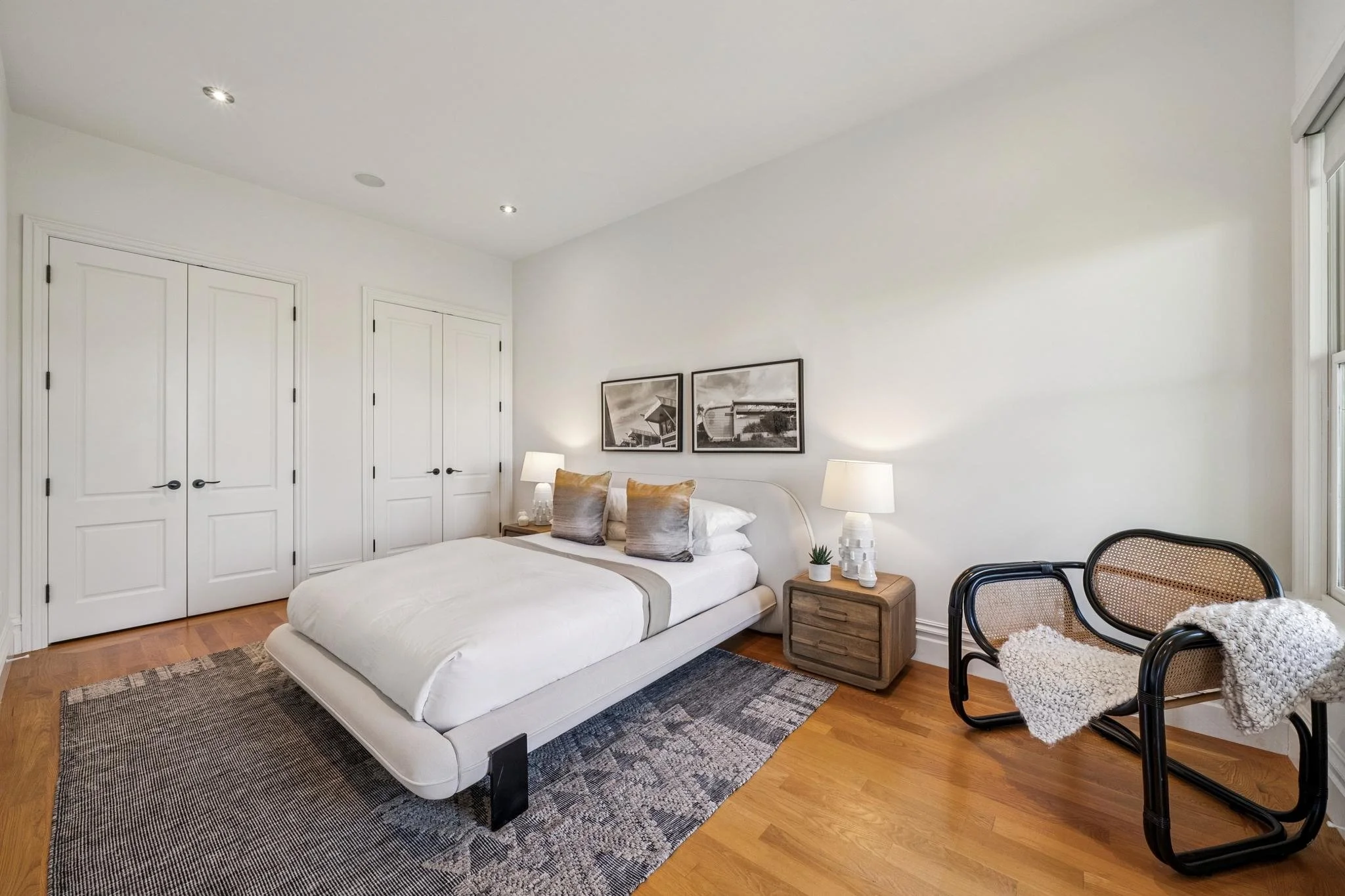 A bedroom featuring a white bed with pillows, a wooden nightstand with lamps and decoration, framed pictures on the wall, and black chairs with woven seats near a window.