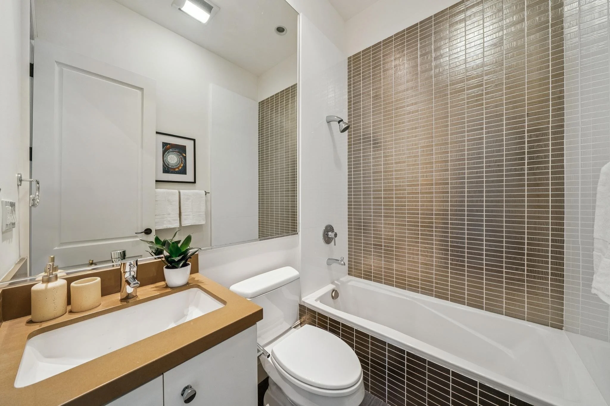 Modern bathroom with a bathtub, a toilet, a sink with a beige countertop, a mirror, framed art, a small plant, and dark brown tile accents.