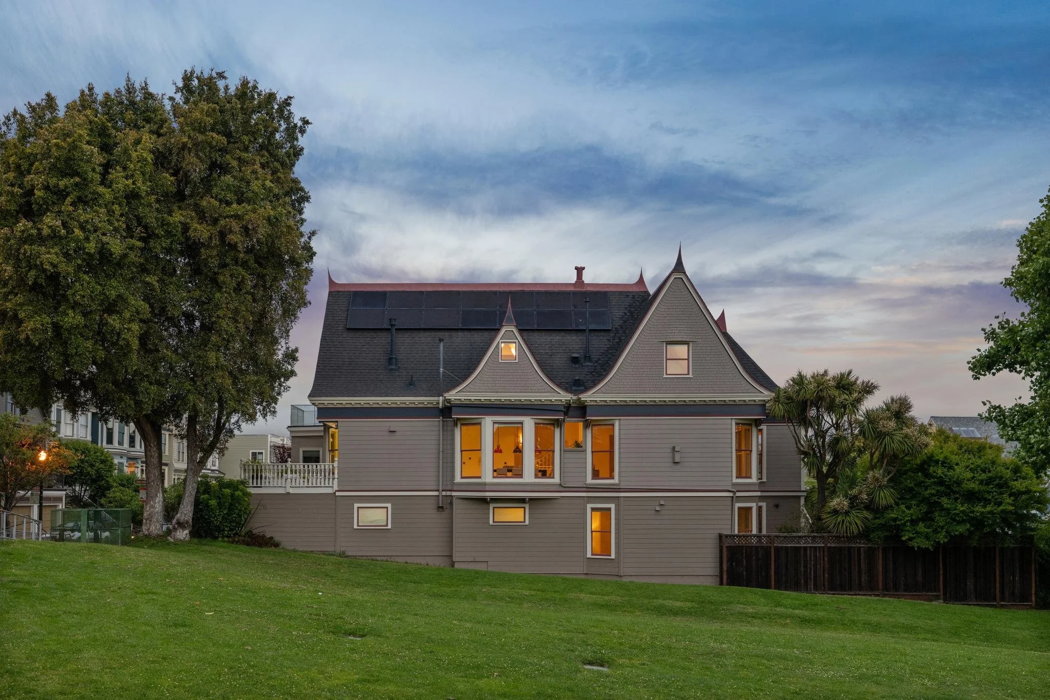 A large house with a triangular roof has solar panels and several illuminated windows, surrounded by tall trees and a well-maintained green lawn at dusk.