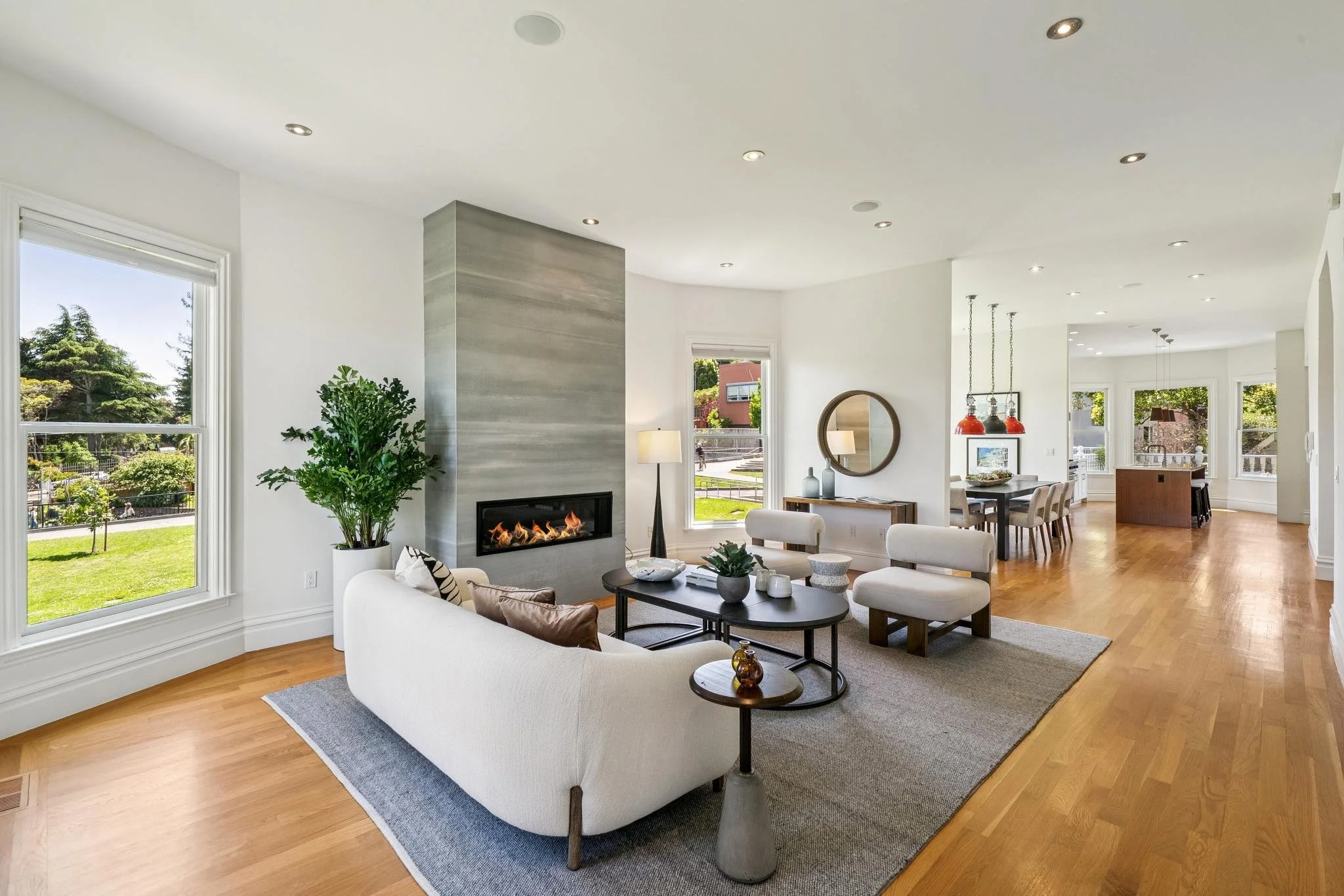 Bright, modern living room with white walls, large windows, hardwood floors, and a fireplace with a gray stone surround. The room is furnished with white sofas, a black coffee table, and decorative items. A large leafy plant is near the window, and a