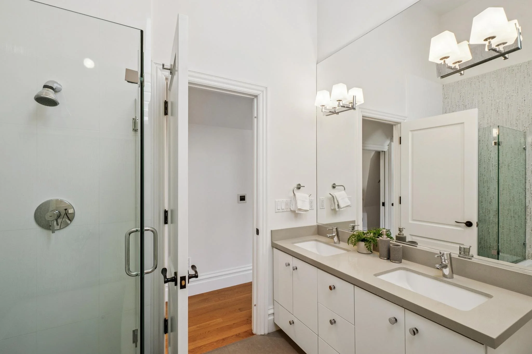 A bathroom with a glass shower on the left, a white vanity with two sinks and a large mirror on the right, and a wall-mounted light fixture with three white shades above the mirror. There are two towels on towel rings next to the vanity, and some pla